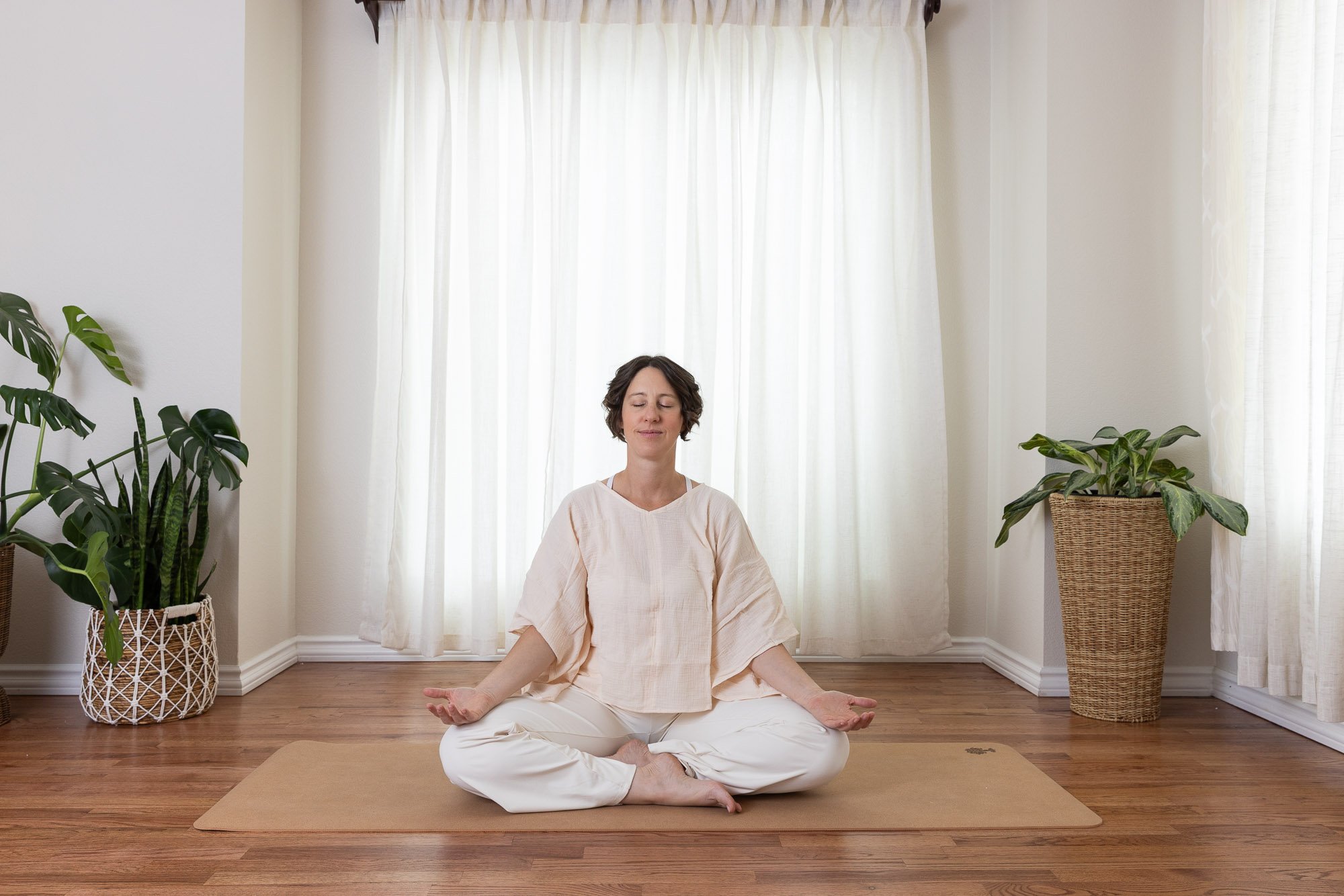 A woman practicing yoga and meditation in a bright living room, seated cross-legged on a yoga mat with her eyes closed, surrounded by potted plants and sheer curtains.