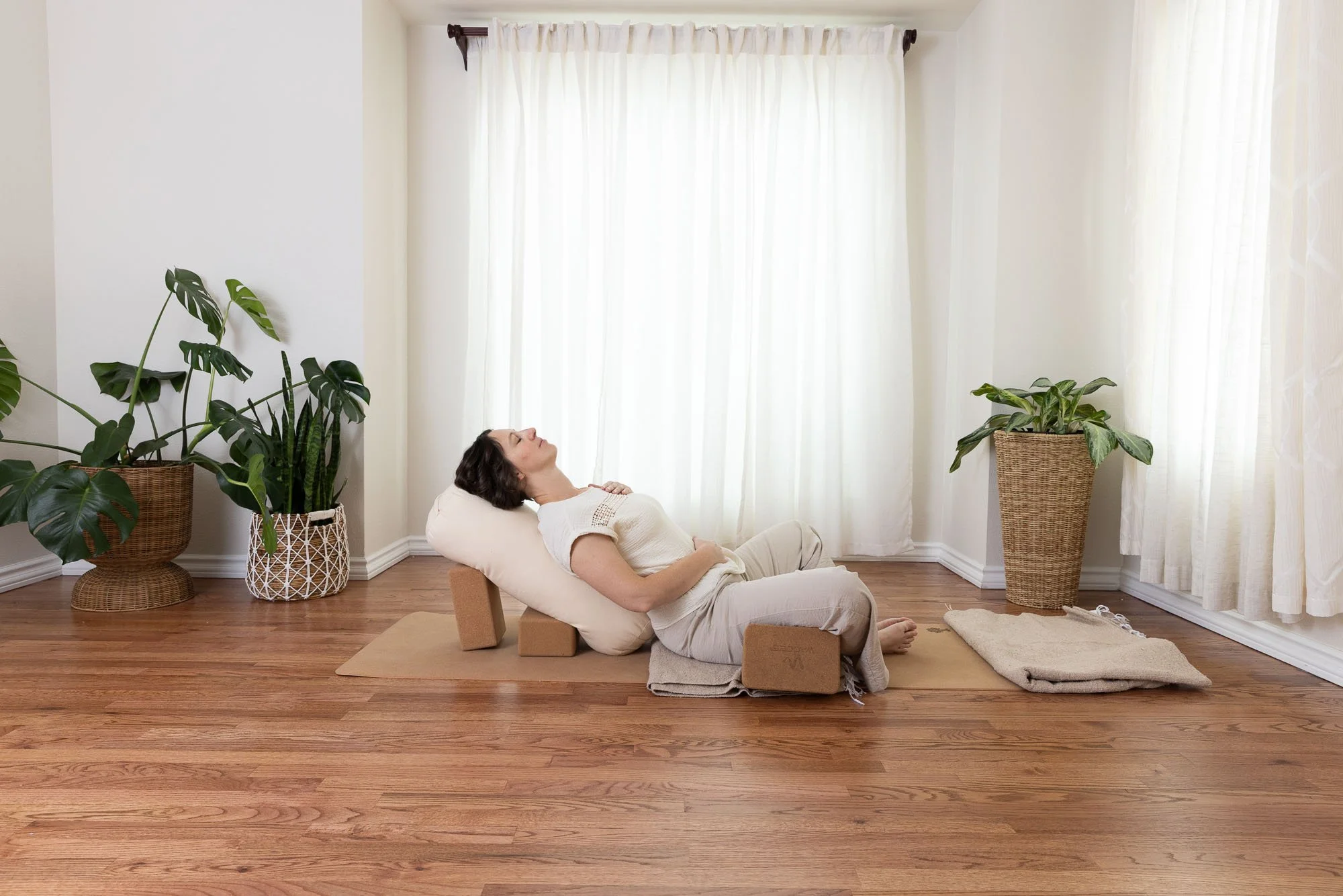 A woman practicing restorative yoga using foam blocks and bolsters in a home setting. She reclines on her back, supported by cushions and yoga props, in front of white curtains with potted plants on either side.