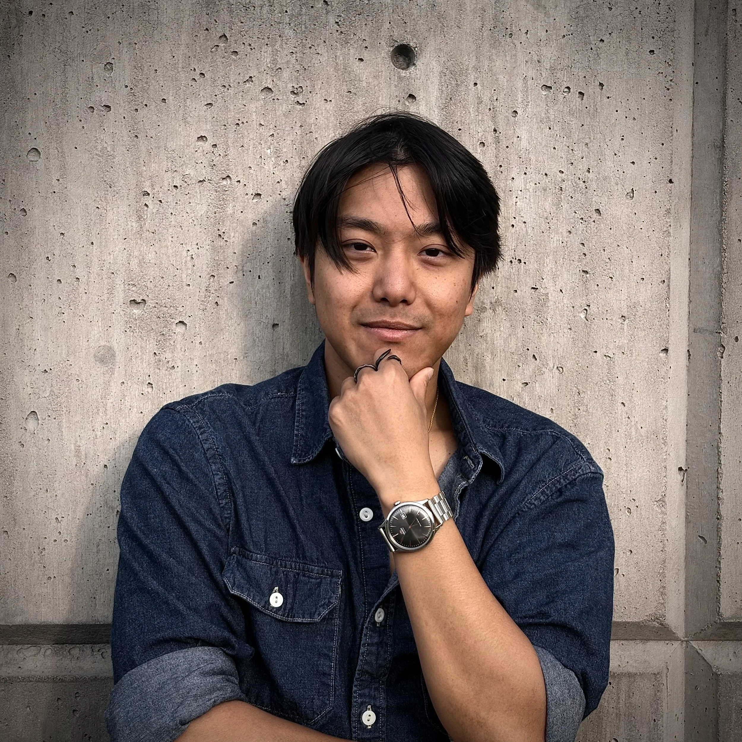 Jeddie in a blue, denim shirt, hand on chin, leaning against a concrete wall.