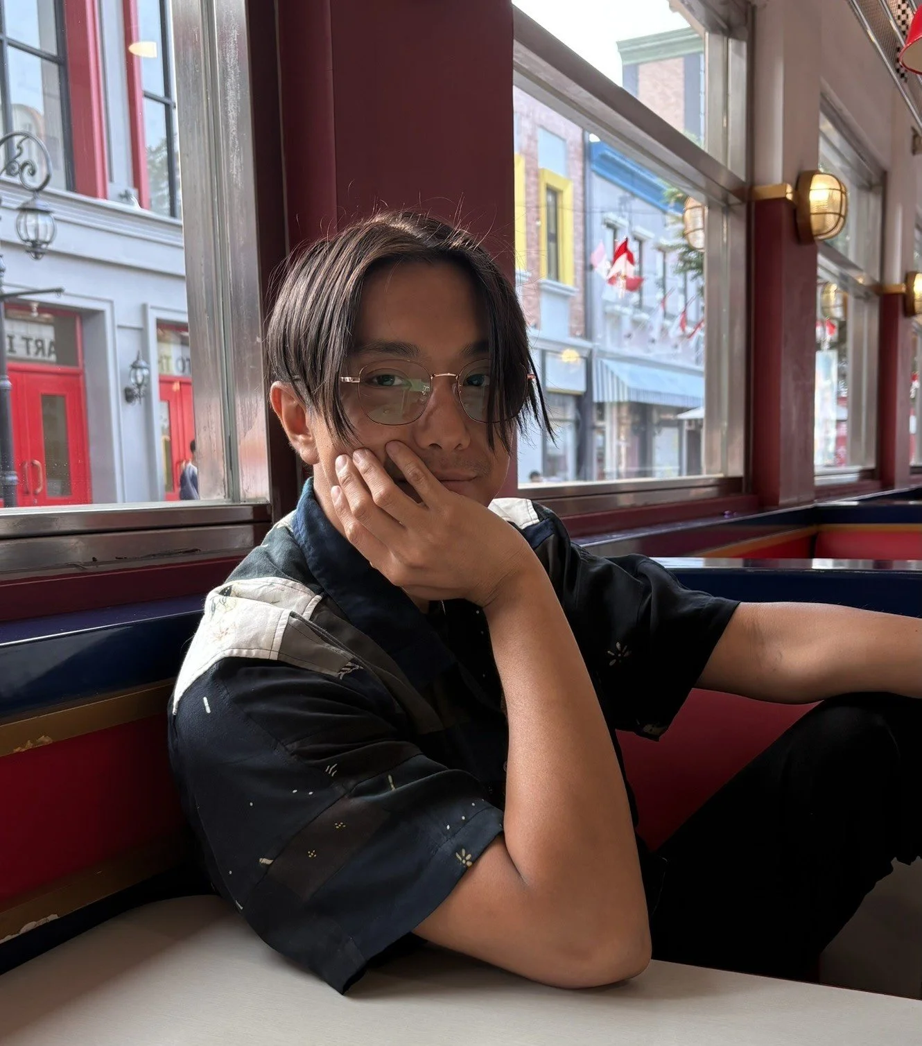 Jeddie seated at a diner, wearing a colorful, patterned-shirt and eyeglasses. Jeddie's smiling face is covered by his right hand.
