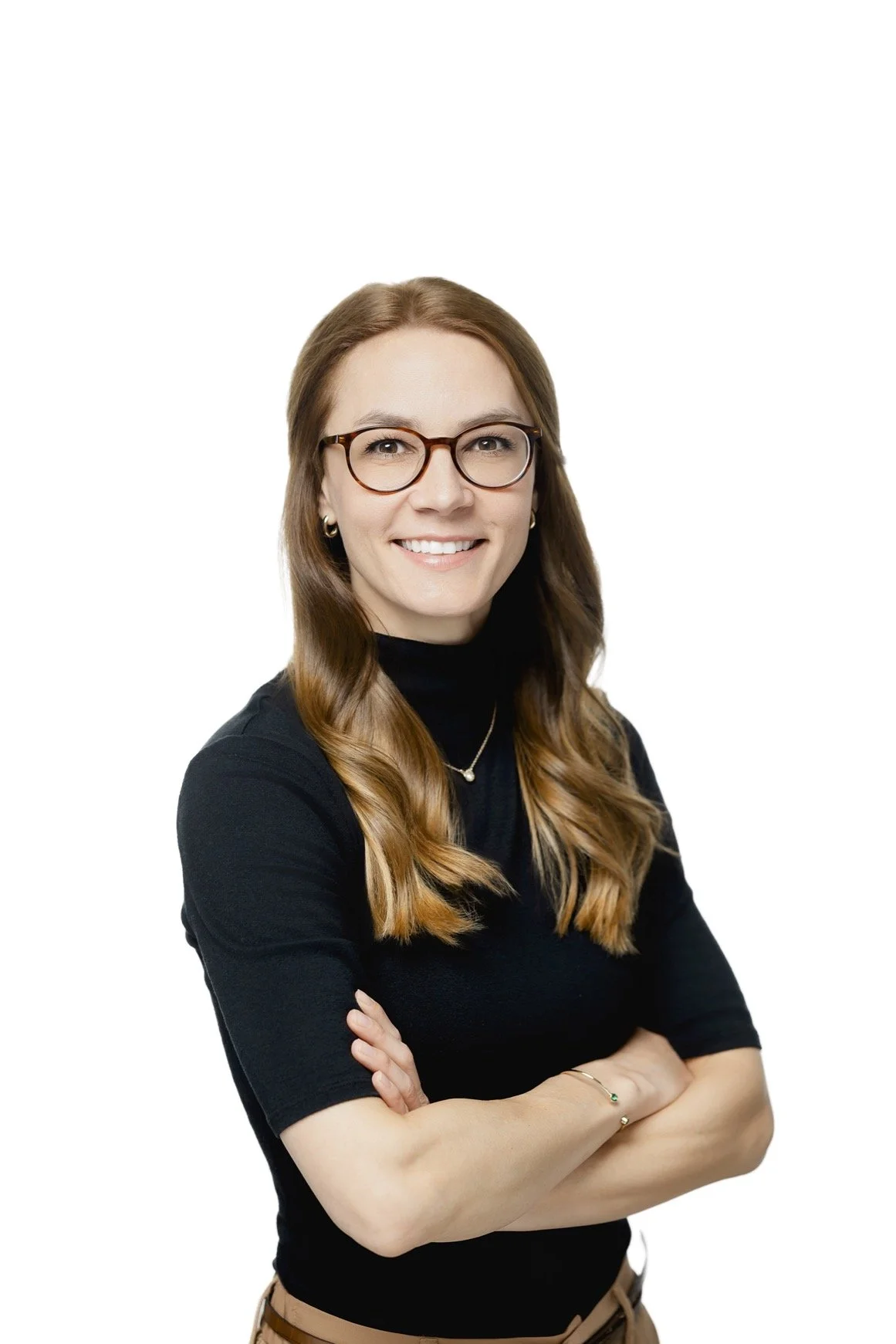 Portrait of a woman with long, wavy brown hair wearing glasses, a black turtleneck, and gold jewelry, smiling with crossed arms against a white background.