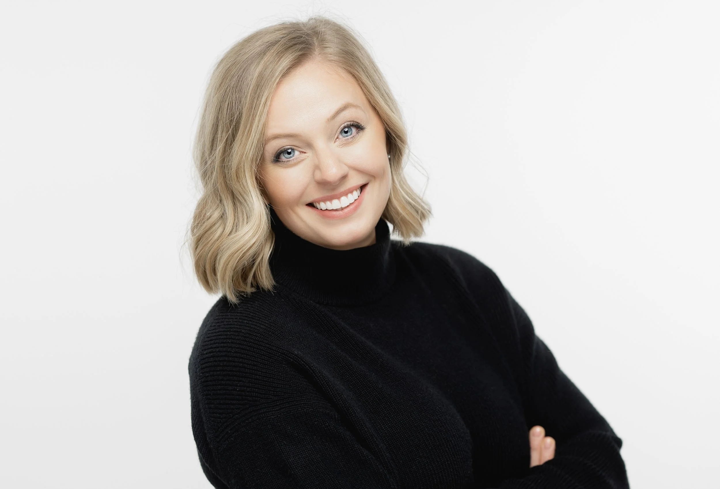 A woman with blonde hair in loose waves, wearing a black turtleneck sweater, smiling against a white background.