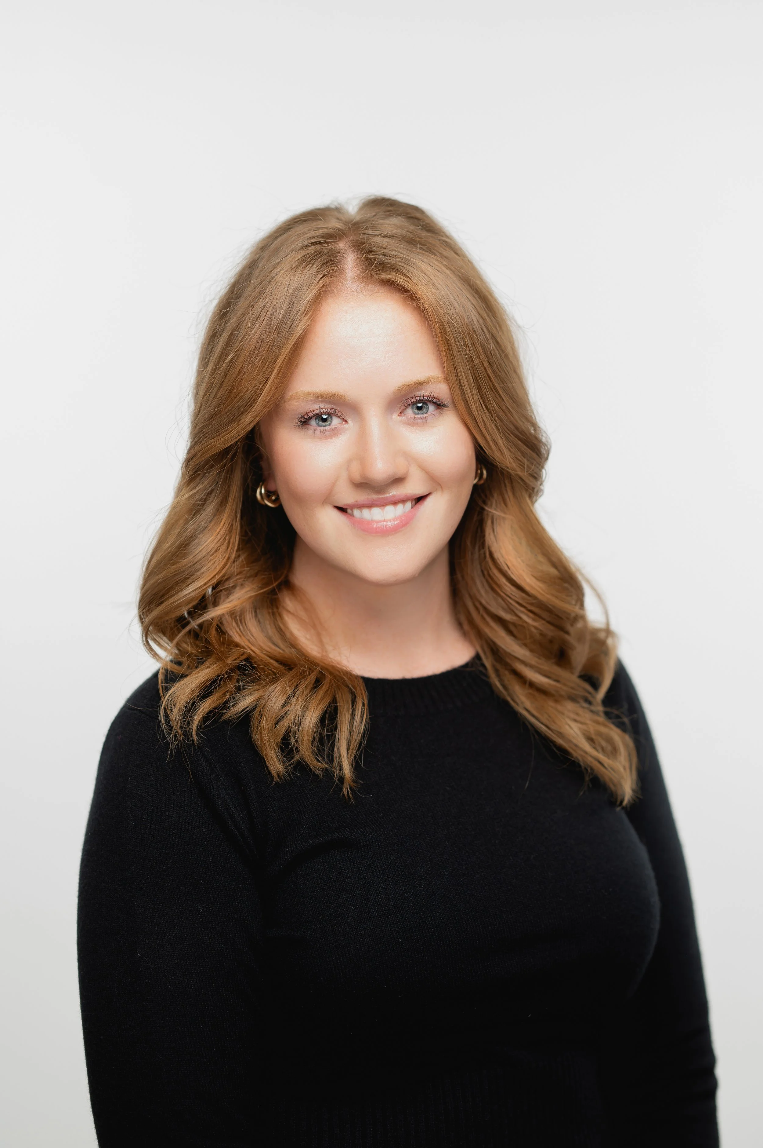 A woman with light skin and long wavy auburn hair wearing a black top, smiling against a plain white background.