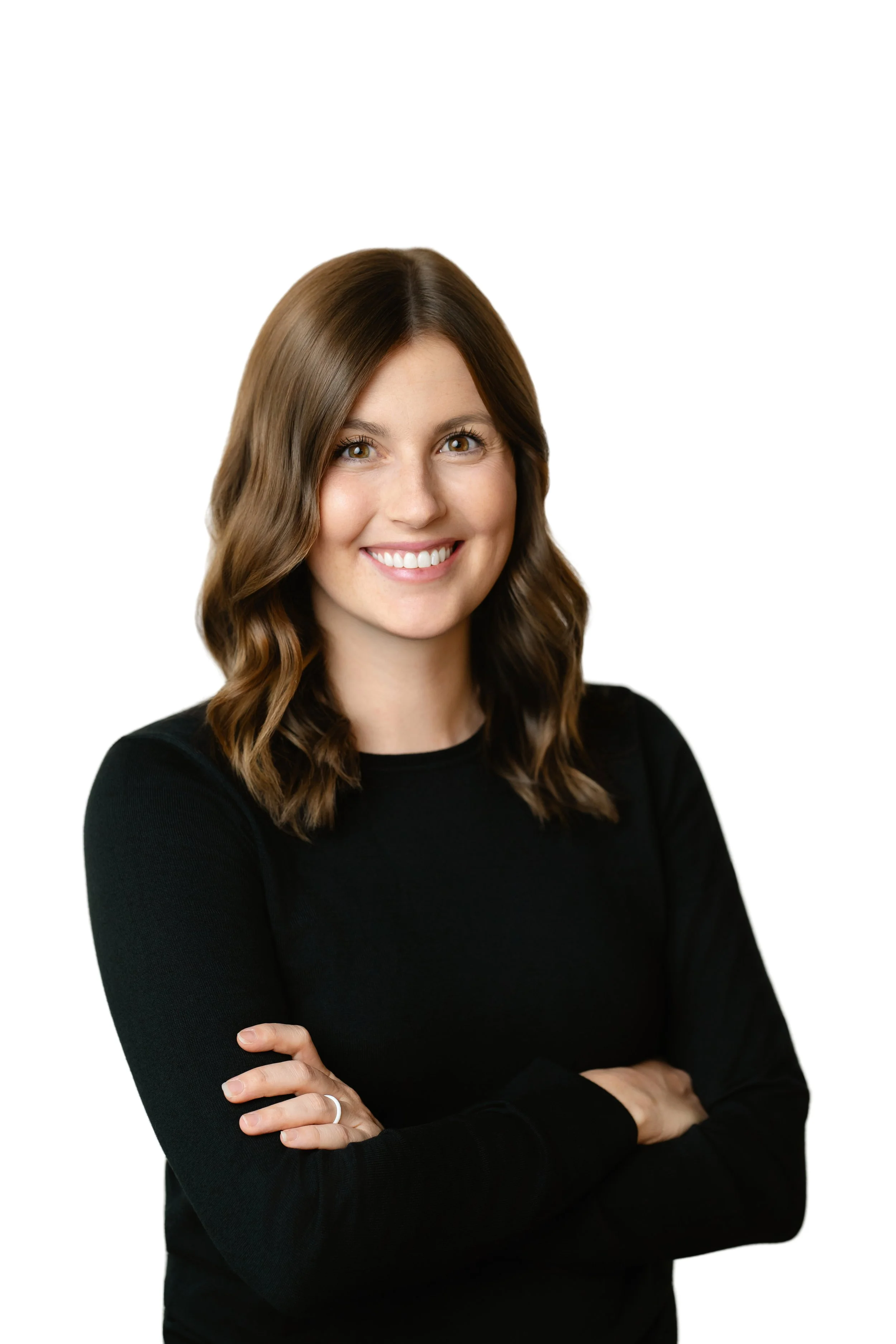 A woman with brown, wavy hair wearing a black long-sleeve top, smiling and crossing her arms, against a plain white background.