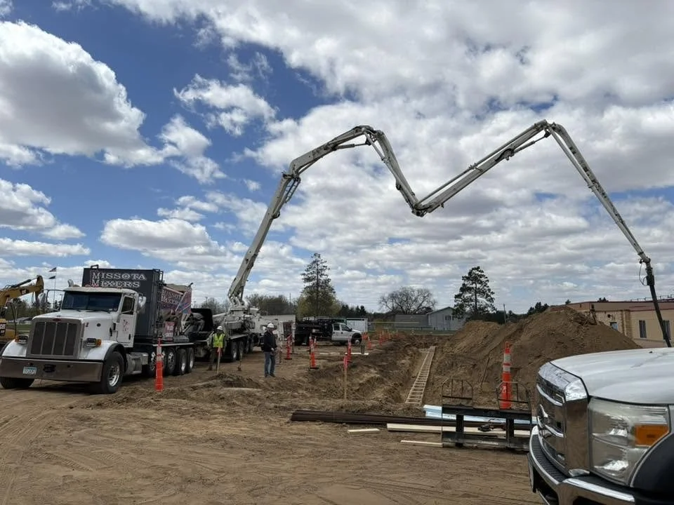 Construction site with a concrete pump truck pouring concrete, workers, dirt mounds, and construction materials under a cloudy sky.