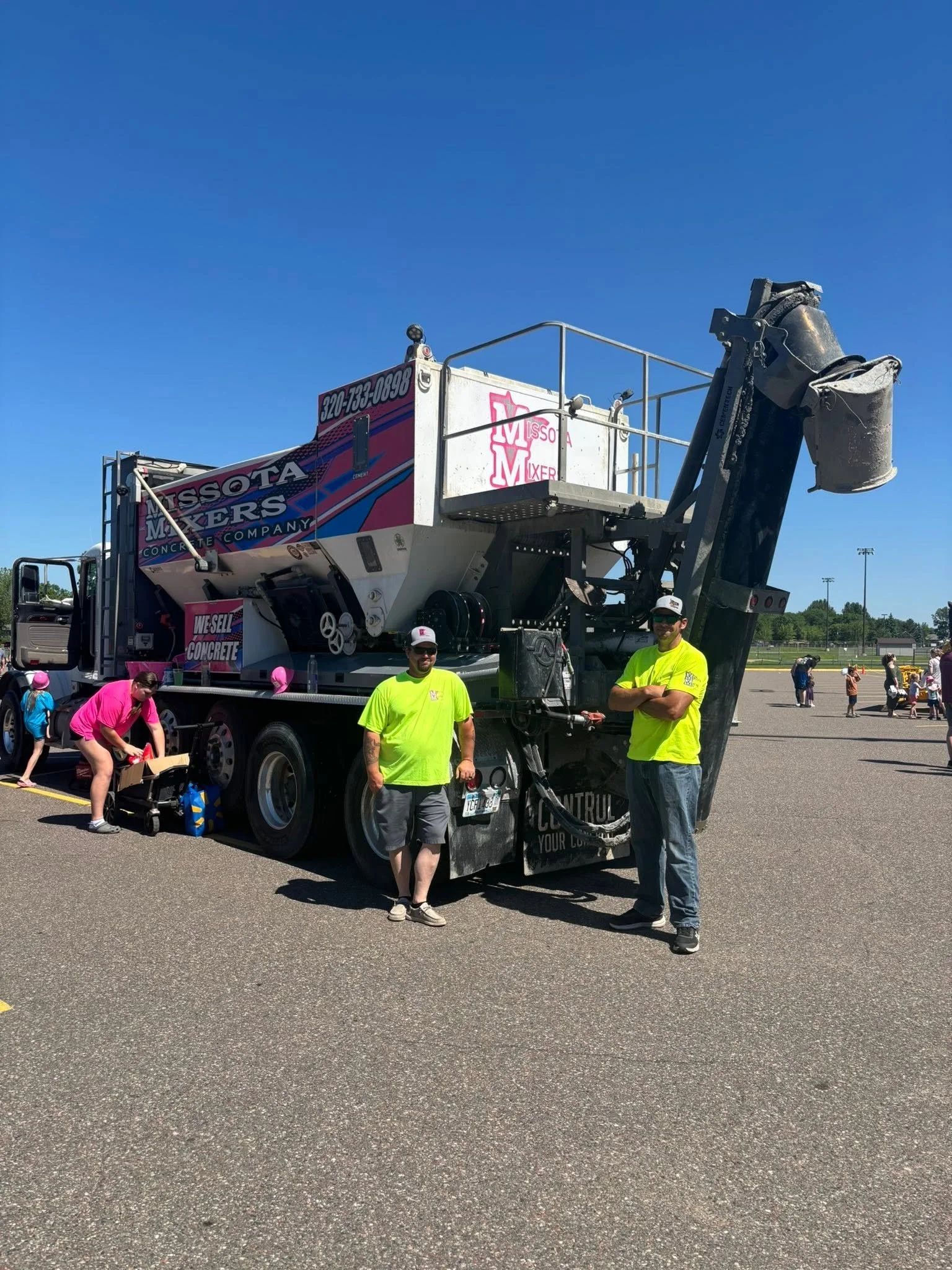 A concrete mixer truck with people in high-visibility yellow shirts standing in front of it in an open parking lot on a sunny day. There are others in the background and some children nearby.