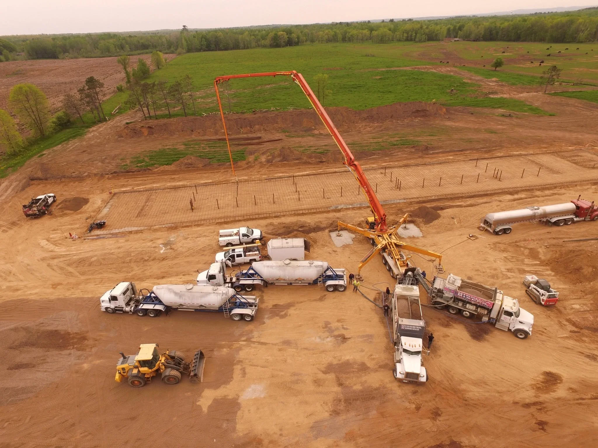 Construction site in a rural area with various construction vehicles and equipment, including cement mixers and trucks, with a large orange concrete pump and rebar mesh for a foundation. Green fields and trees are visible in the background.