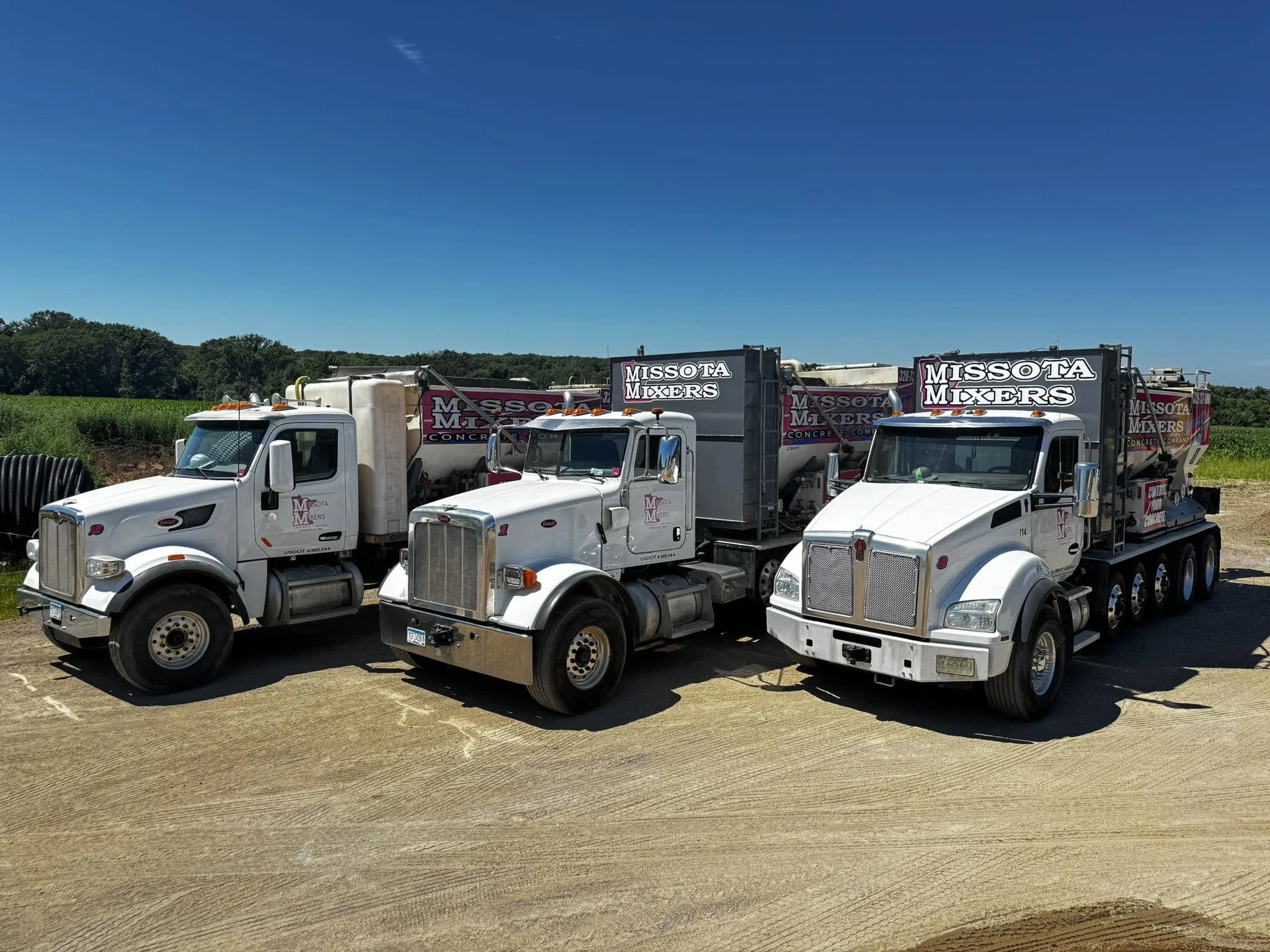 Several white concrete mixer trucks with 'Missota Mixers' logo parked on a dirt field under blue sky with some trees in the background.