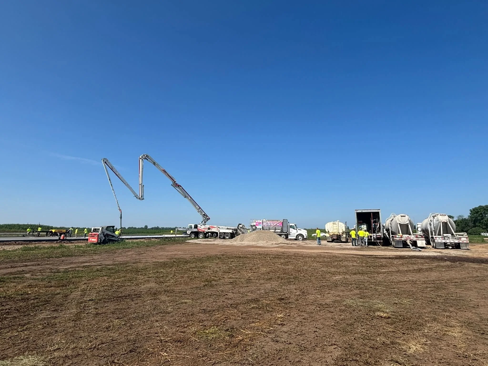 Construction site where workers are pouring concrete on a road. Large concrete pumps are visible, along with trucks and construction equipment under a clear blue sky.