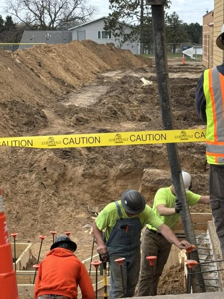 Construction workers in neon shirts and safety gear working on building a foundation, with dug-out earth and caution tape surrounding the site.