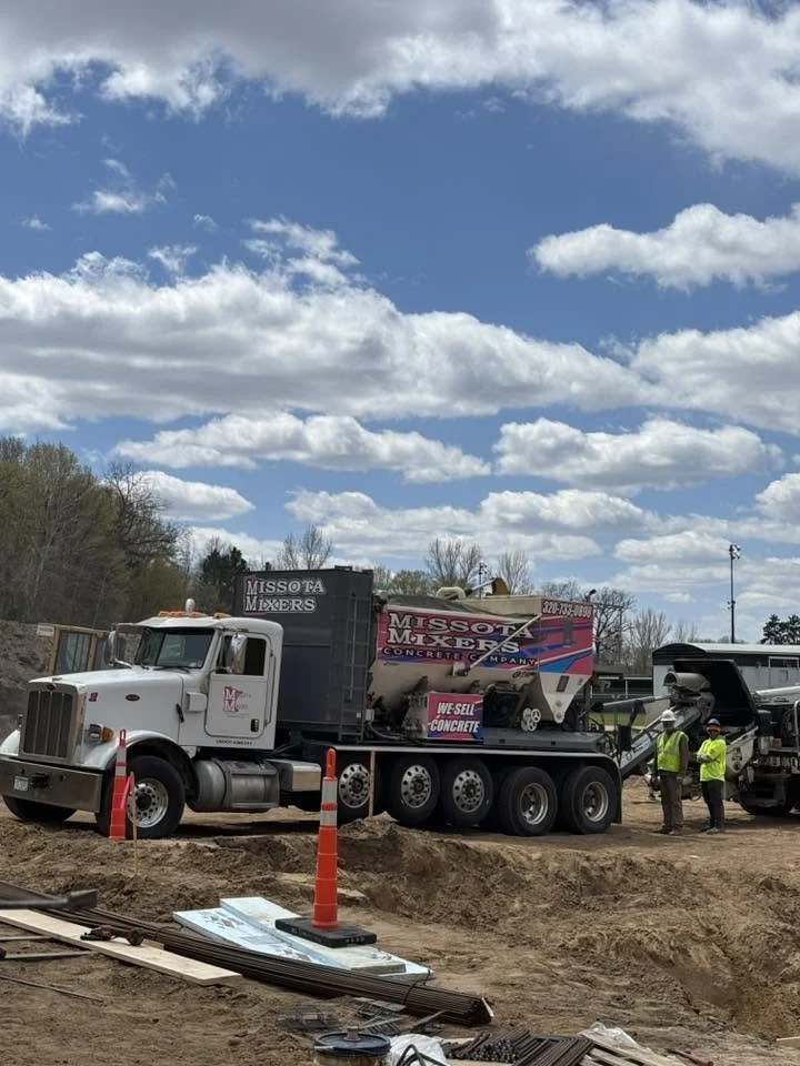 Construction site with a large truck labeled 'Missota Mixers' and 'Concrete Company,' pouring concrete. Workers in safety vests and helmets stand nearby under a blue sky with clouds.