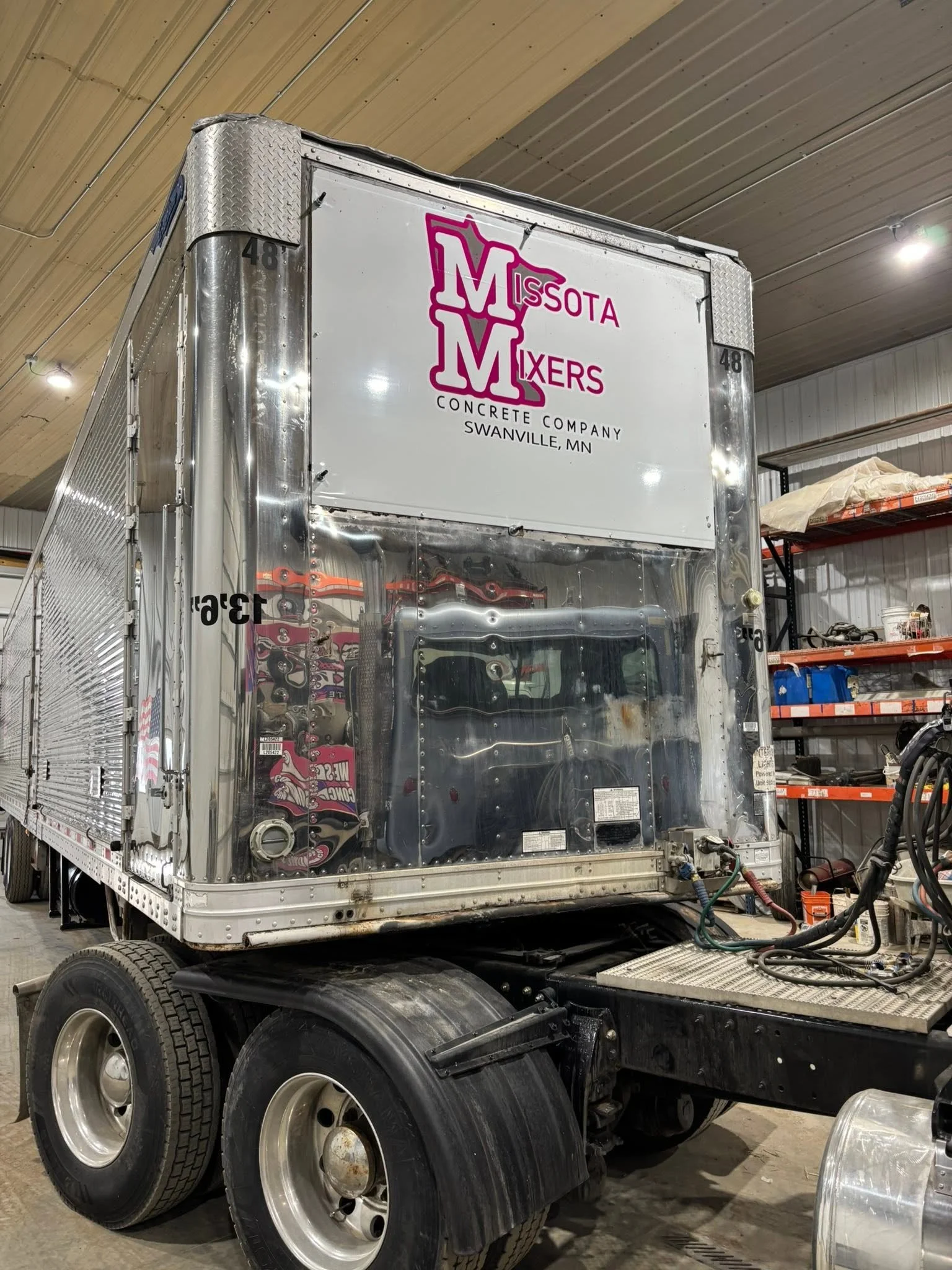 A truck cab is being repaired or built inside a warehouse. The truck has a shiny metal exterior, and there are shelves with tools and materials in the background.