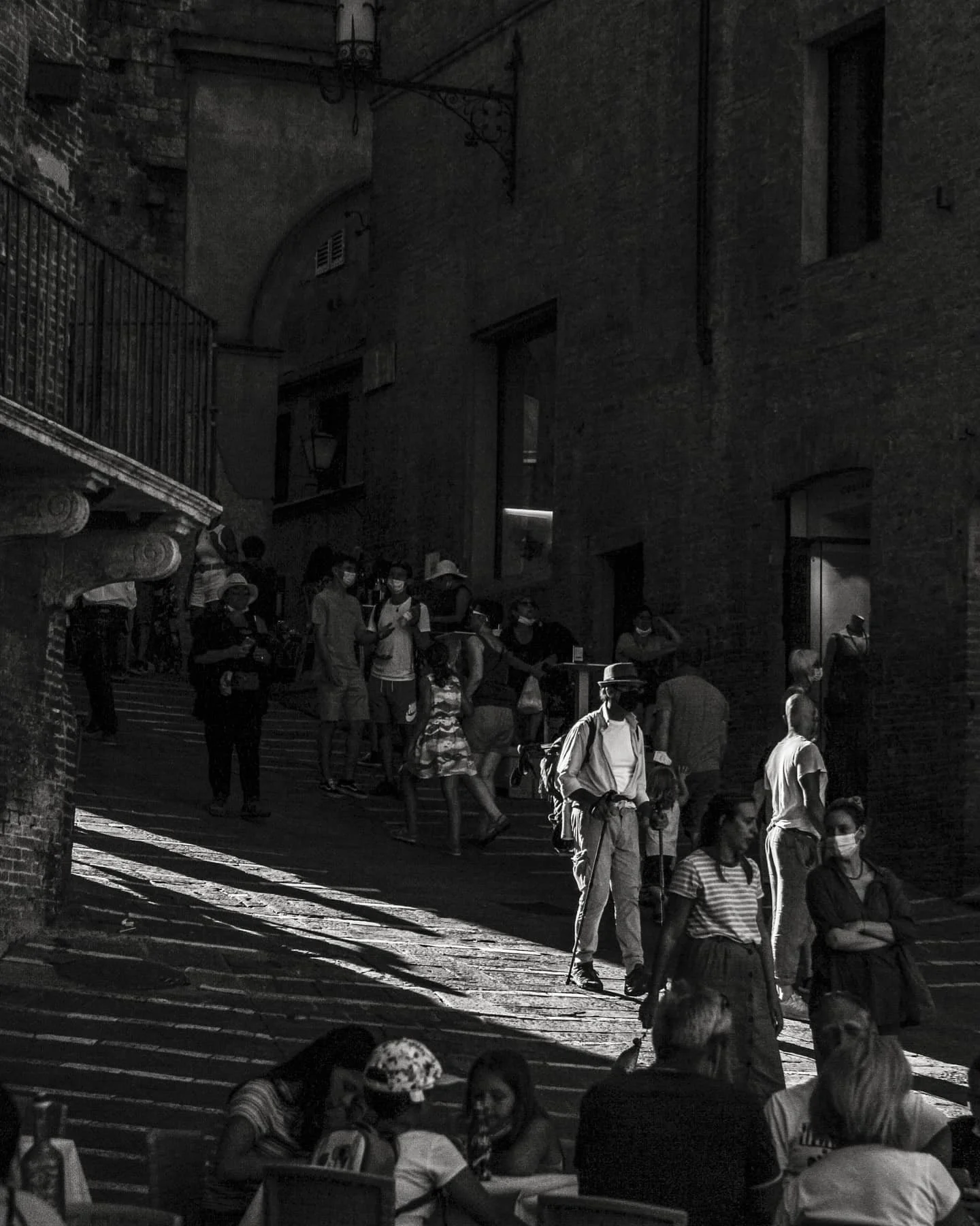 A busy outdoor street scene in black and white with many people walking and sitting at tables. Some are wearing face masks. The background shows building facades, windows, and street lamps with shadows cast on the ground.