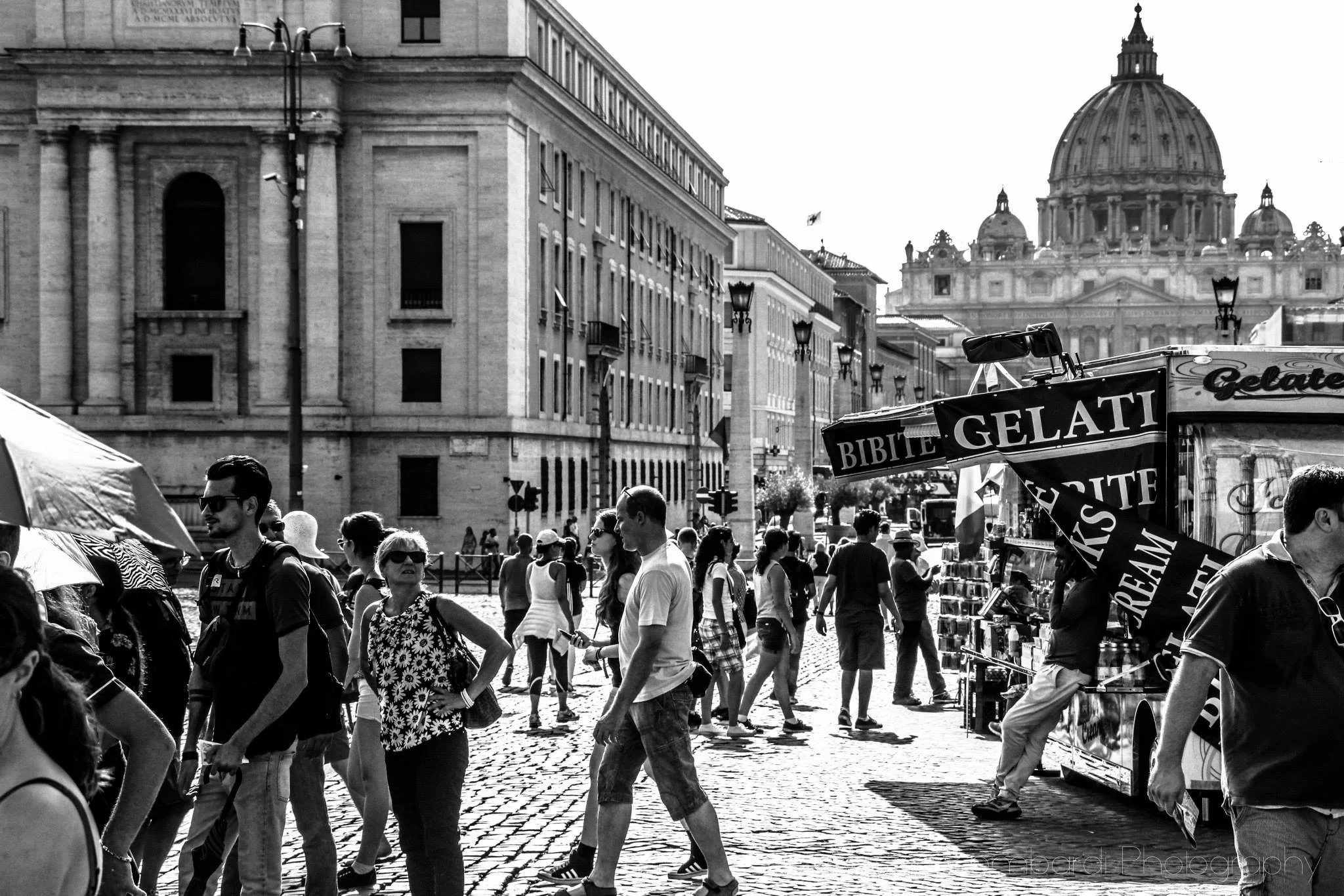 A busy street scene in a European city with crowds of people walking. In the background, there is a large historic building with a dome, and in the foreground, there are street vendors with signs advertising gelato and other treats.