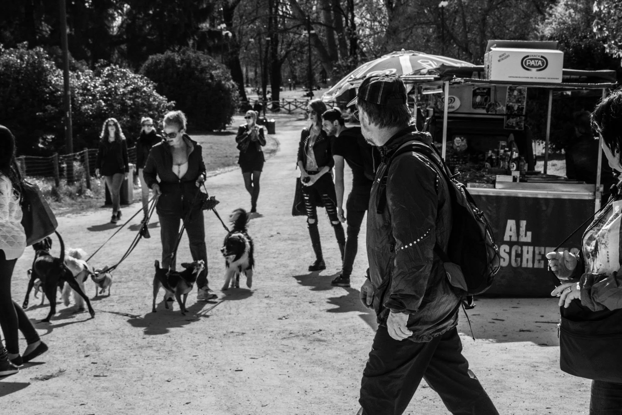 A black and white photo of a group of people in a park, some walking dogs on leashes and others standing, with a food cart in the background.