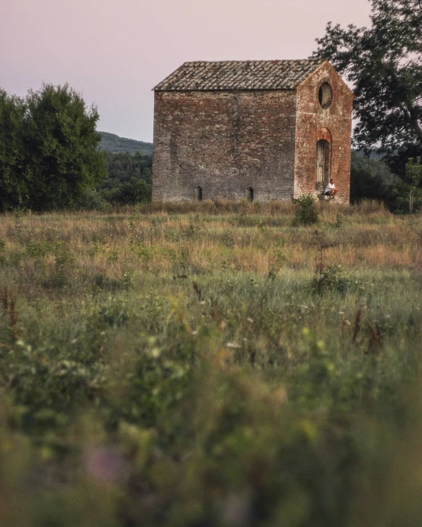 Old brick building with a small circular window, overgrown grassy foreground, trees, and hills in the background during sunset.