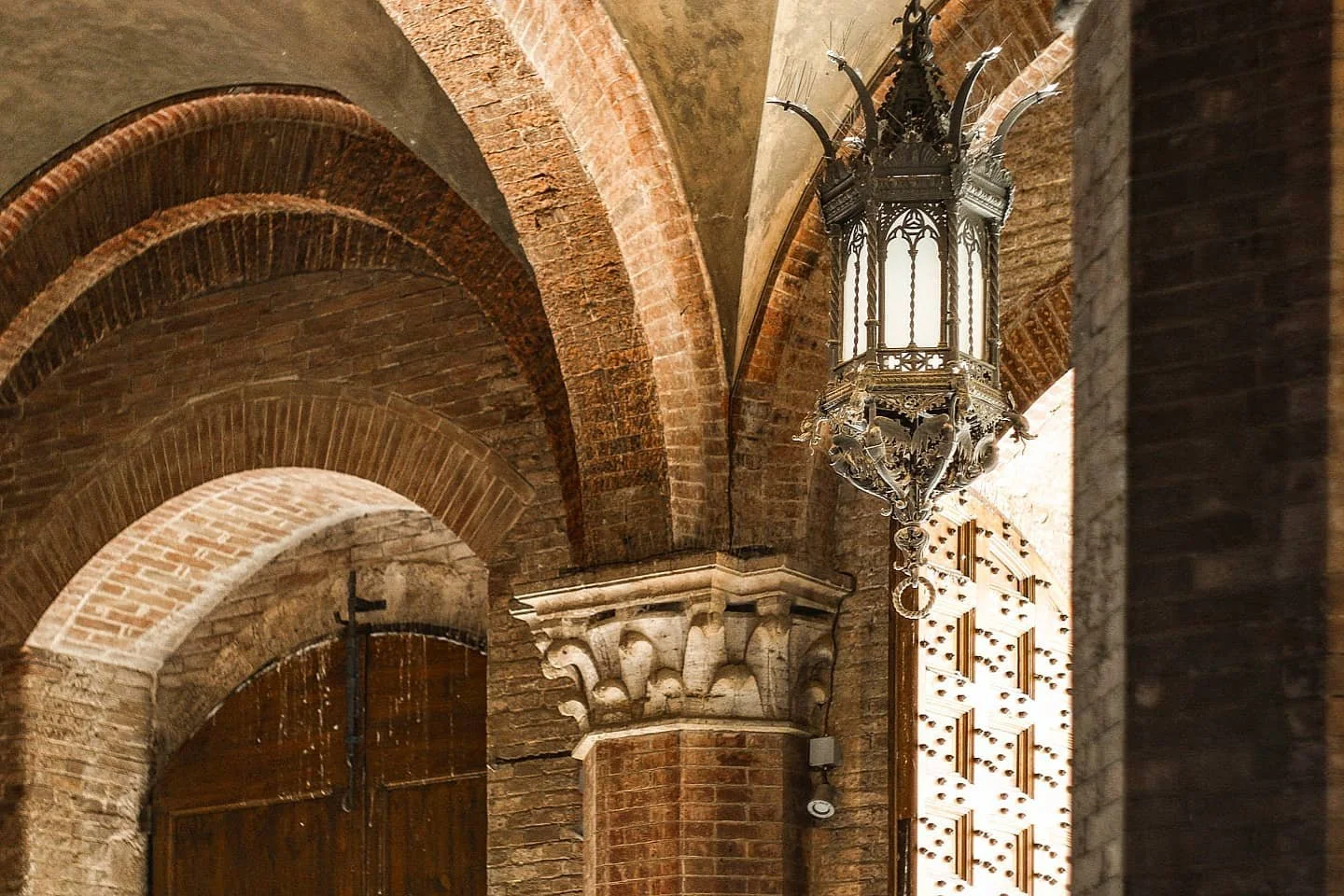 Interior of a brick building featuring arches, a decorative metal lantern hanging from the ceiling, and a wooden door with a cross-shaped handle.