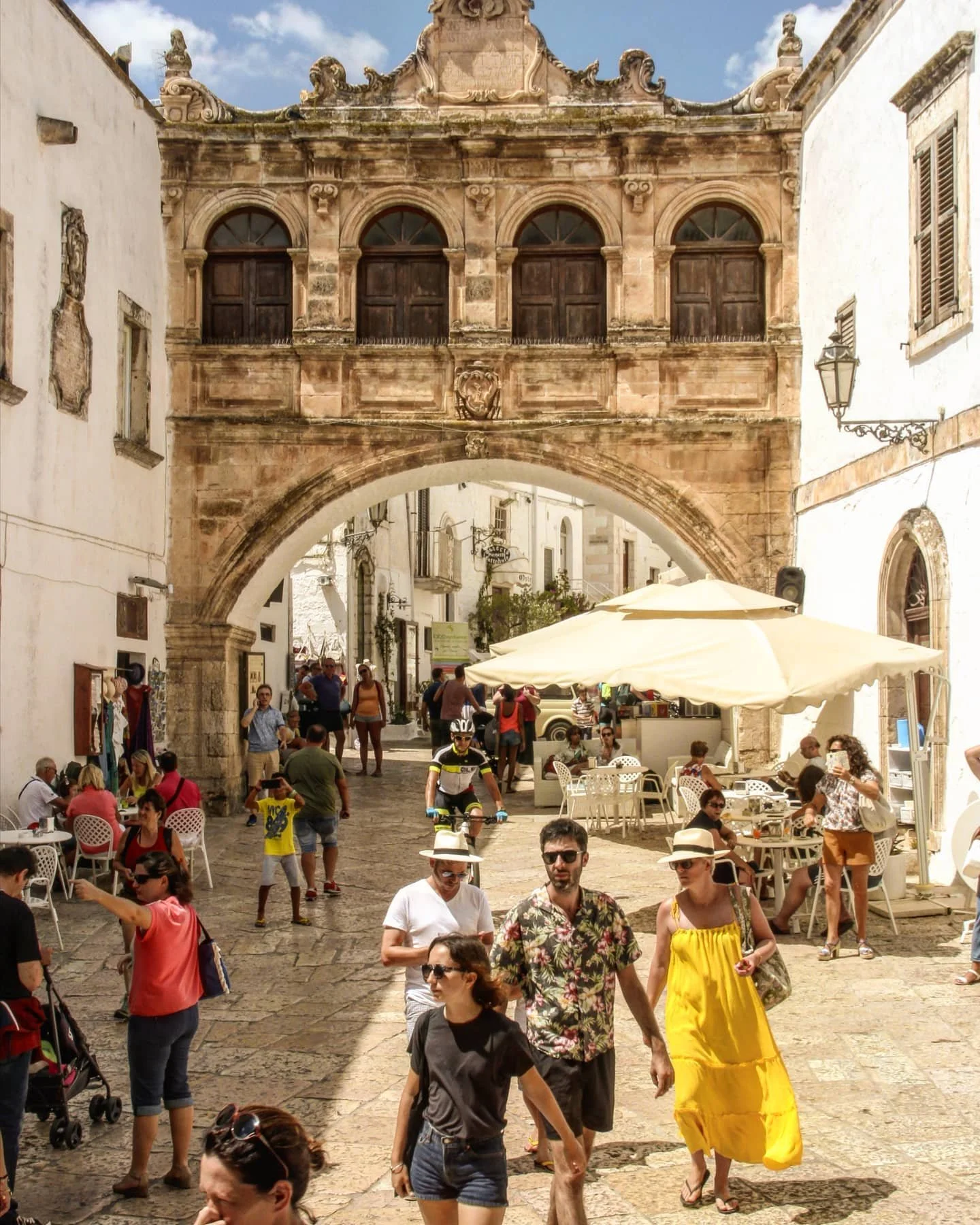 A busy street scene with people walking, sitting at outdoor tables under umbrellas, and a historic archway with detailed stone architecture overhead in a sunny, outdoor marketplace or plaza.