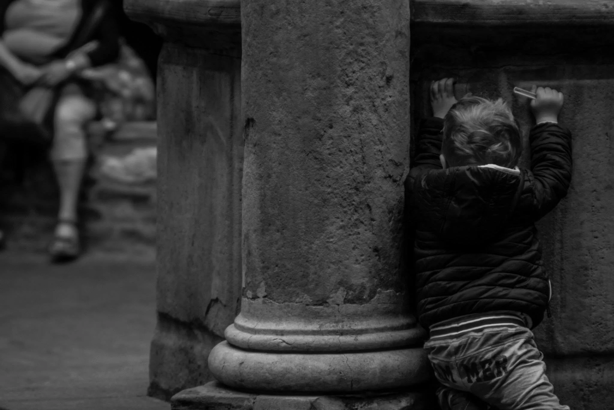 Child crouching behind a stone column, peeking out and holding a pencil, with another child running past in the background, captured in black and white.