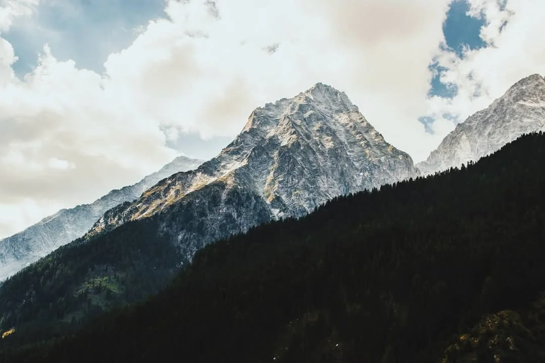 Snow-capped mountain peaks with a forested hillside in the foreground and scattered clouds in the sky.