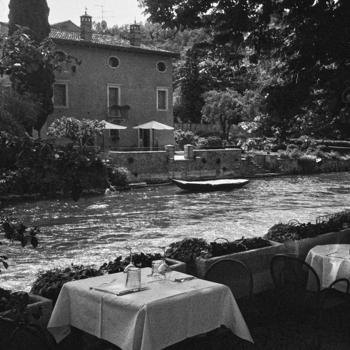 Black and white photo of a riverside restaurant with tables set with napkins and water glasses, overlooking a calm river with a small boat. In the background, there is a house with two umbrellas on its patio, surrounded by trees and some chimneys on rooftops.