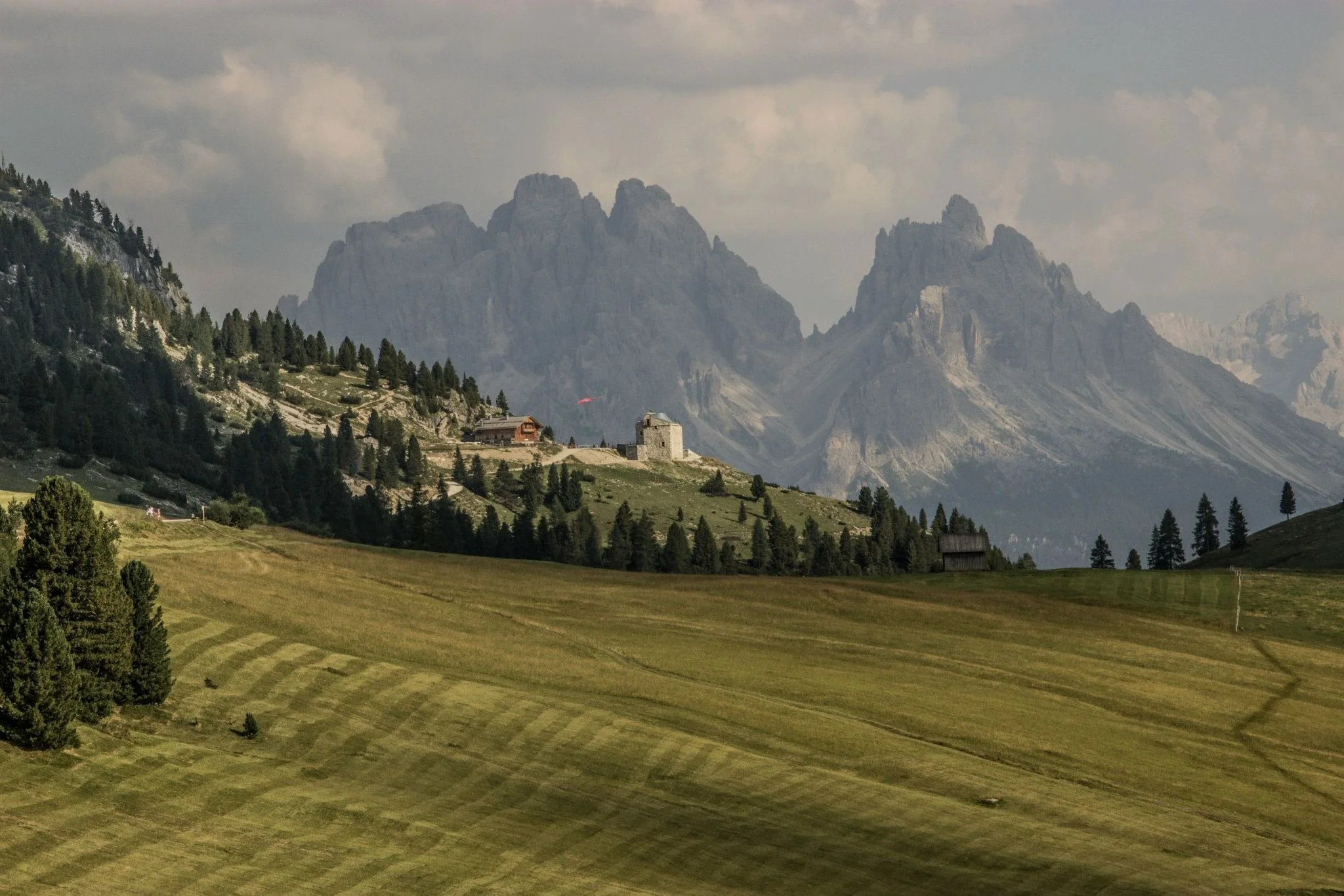 A scenic mountain landscape with rocky peaks in the background and a green, hilly foreground dotted with trees and small buildings.