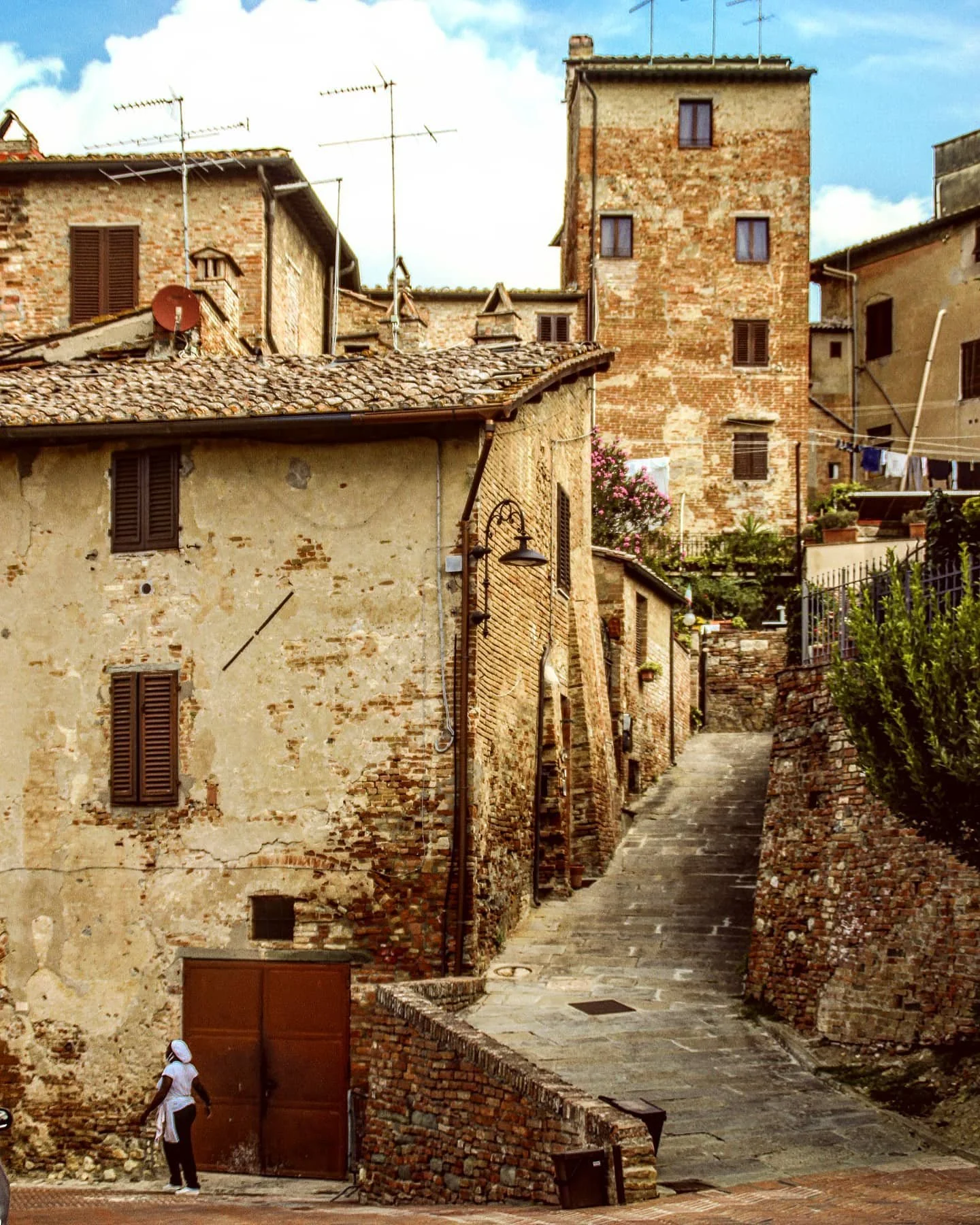 A narrow stone street in an old European village, with rustic brick and stucco buildings, some with wooden shutters, laundry hanging, and lush greenery, under a partly cloudy sky.