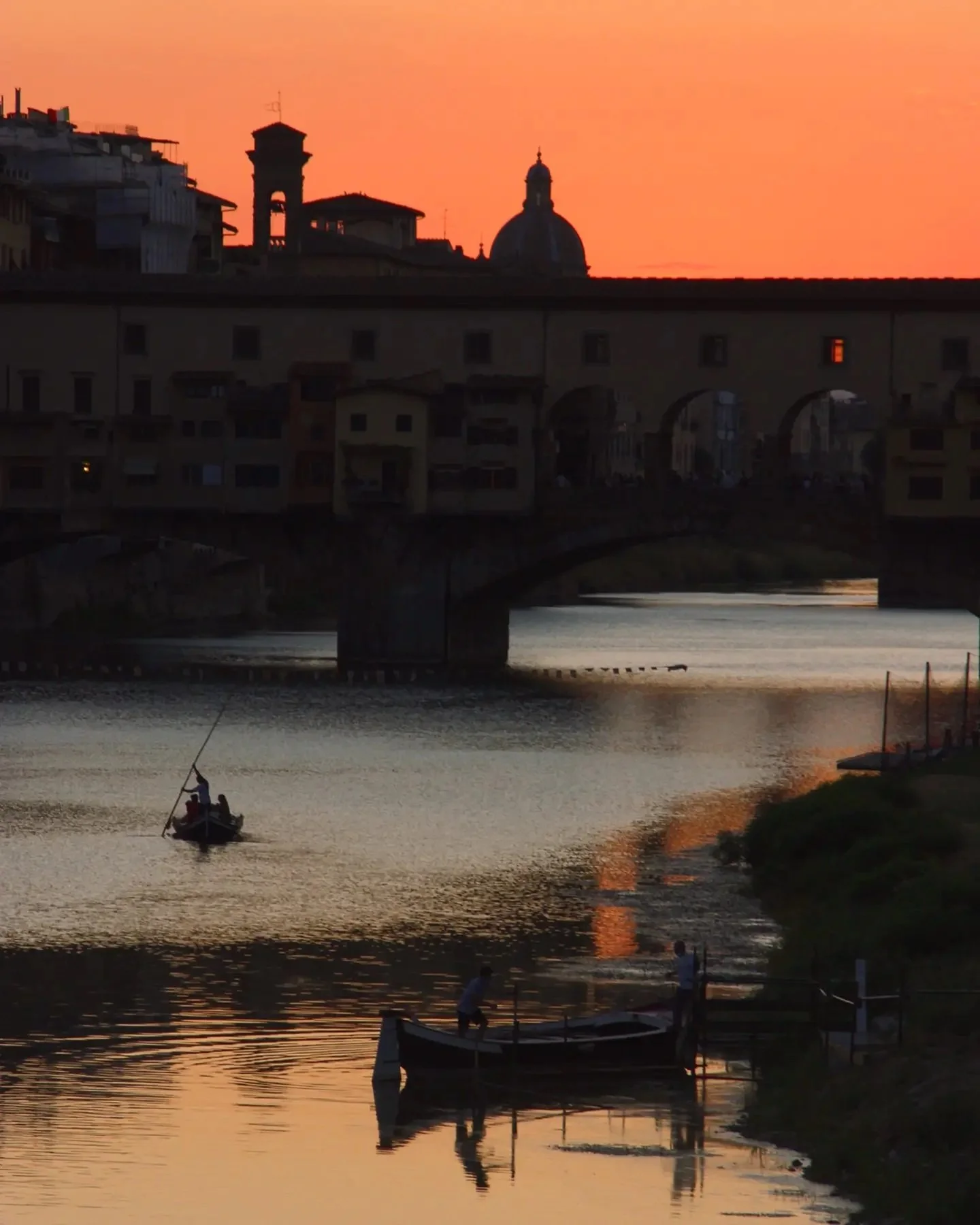 A sunset scene over a river with a small boat and two people fishing, buildings and a bridge with arches in the background, and a warm orange sky.