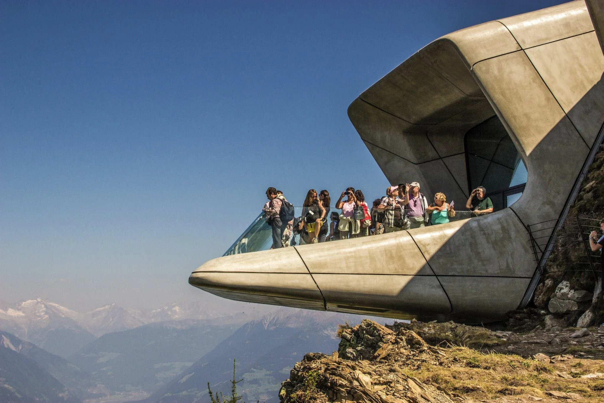 Group of tourists standing on an observation deck with a modern architectural structure, overlooking mountains and a valley during daytime.