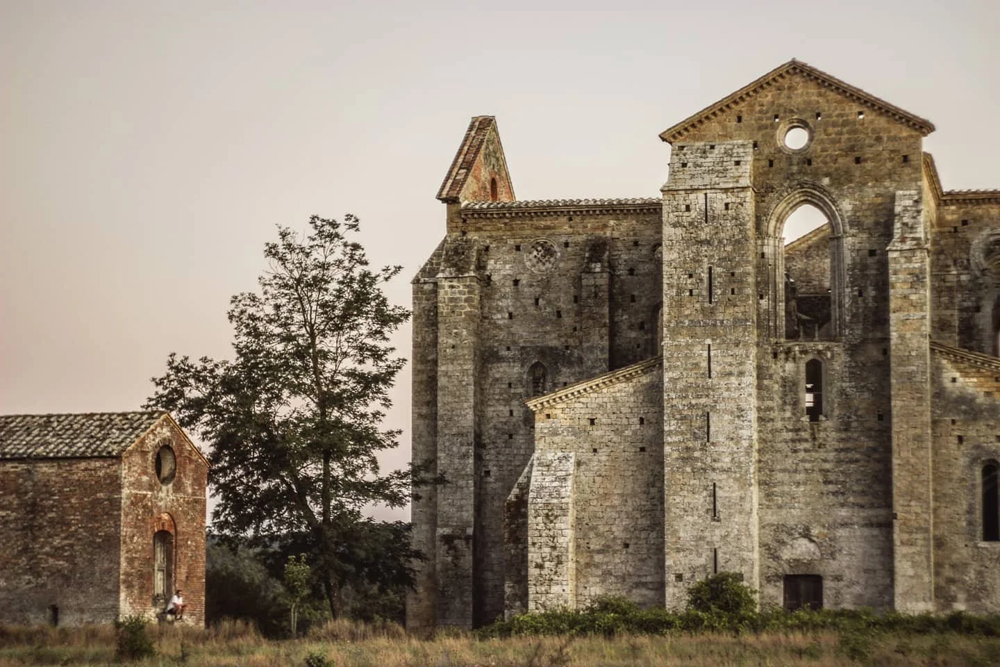 Ancient stone church ruins with tall, weathered walls, arched windows, and a triangular roof, surrounded by grass and trees.