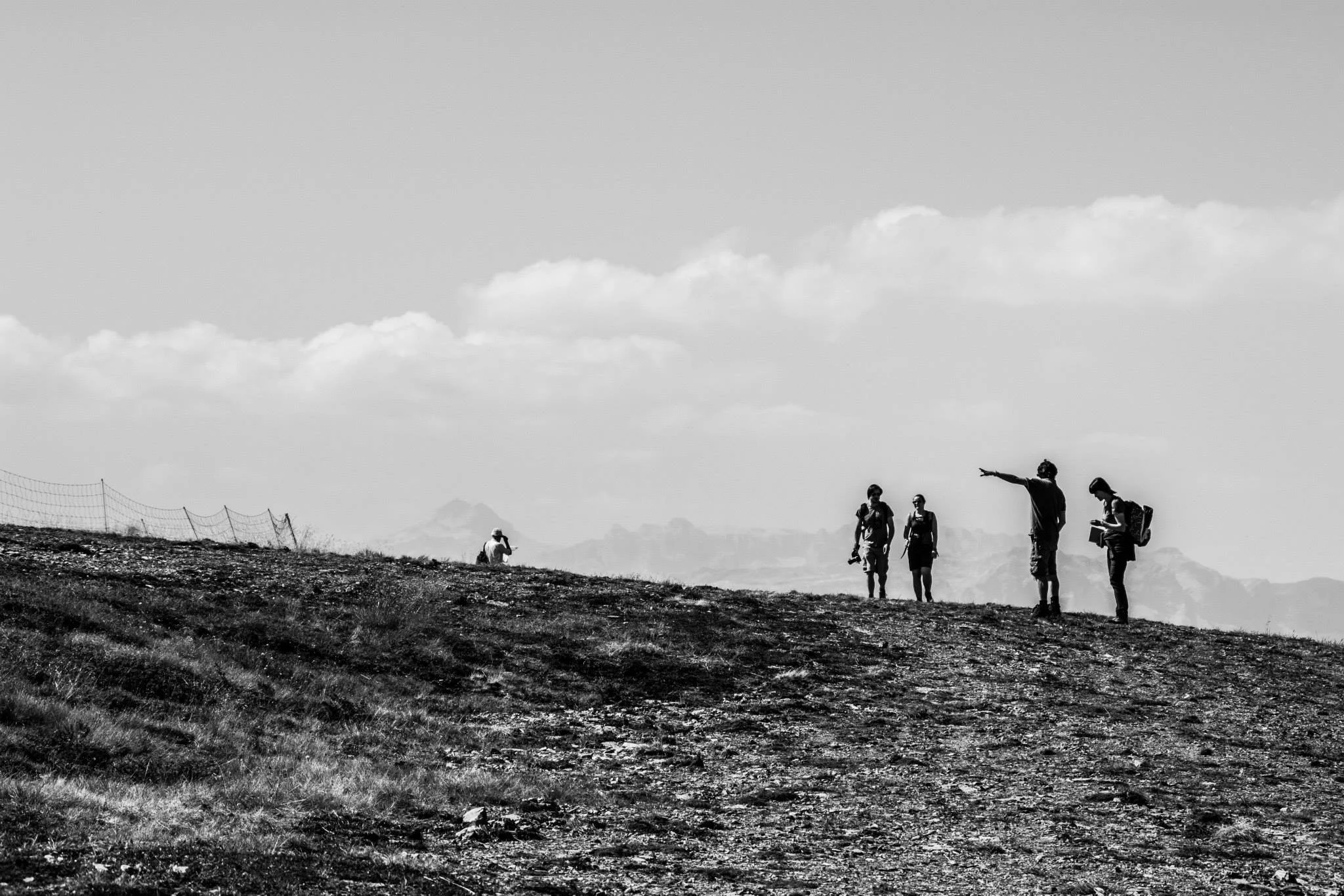 Black and white photo of a group of casually dressed people walking outdoors on a grassy hilltop under a cloudy sky. One person is pointing ahead, and others are carrying backpacks and cameras.