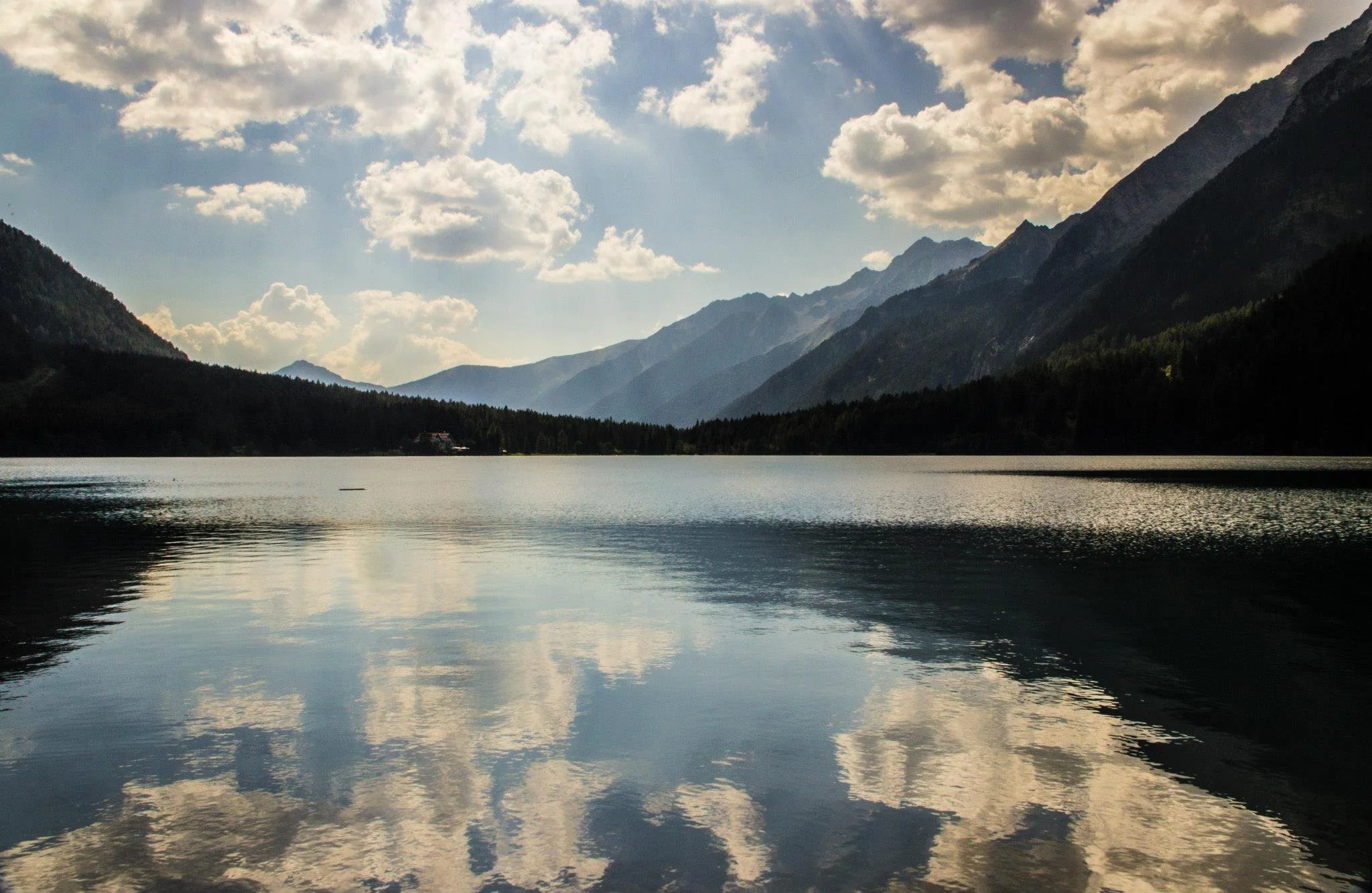 A tranquil mountain lake reflecting the cloudy sky and surrounding mountains with lush green trees along the shoreline.