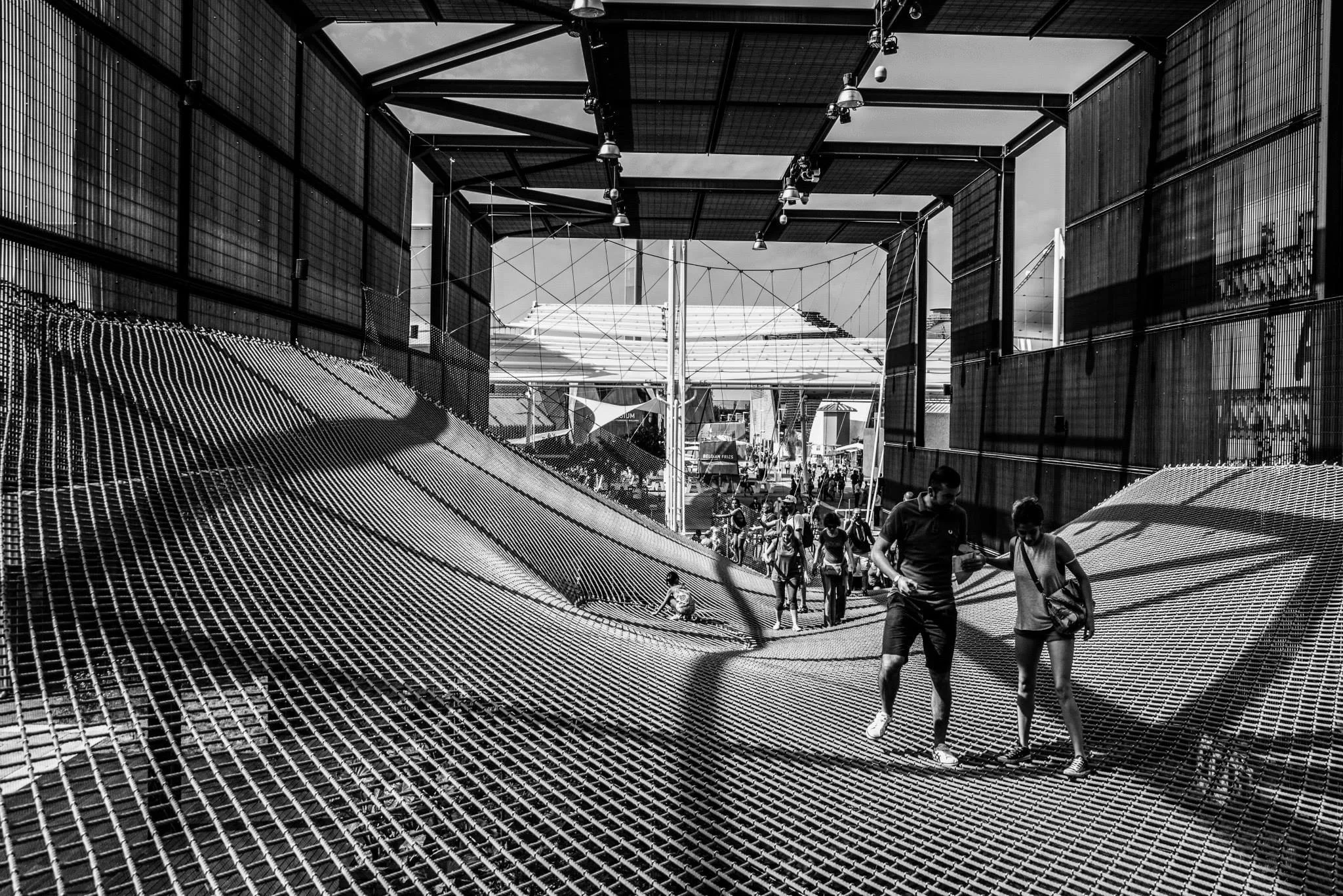 People walking on a metallic grid walkway under a modern architectural structure with a glass and metal canopy.
