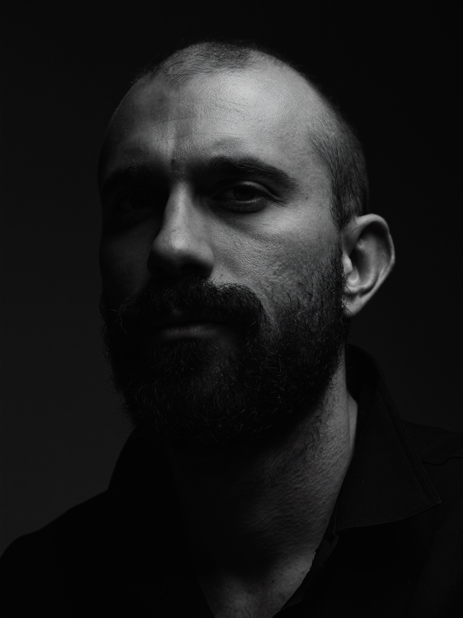 Black and white close-up portrait of a man with a beard and mustache, looking slightly to the side, with a serious expression, dark background, high contrast lighting.