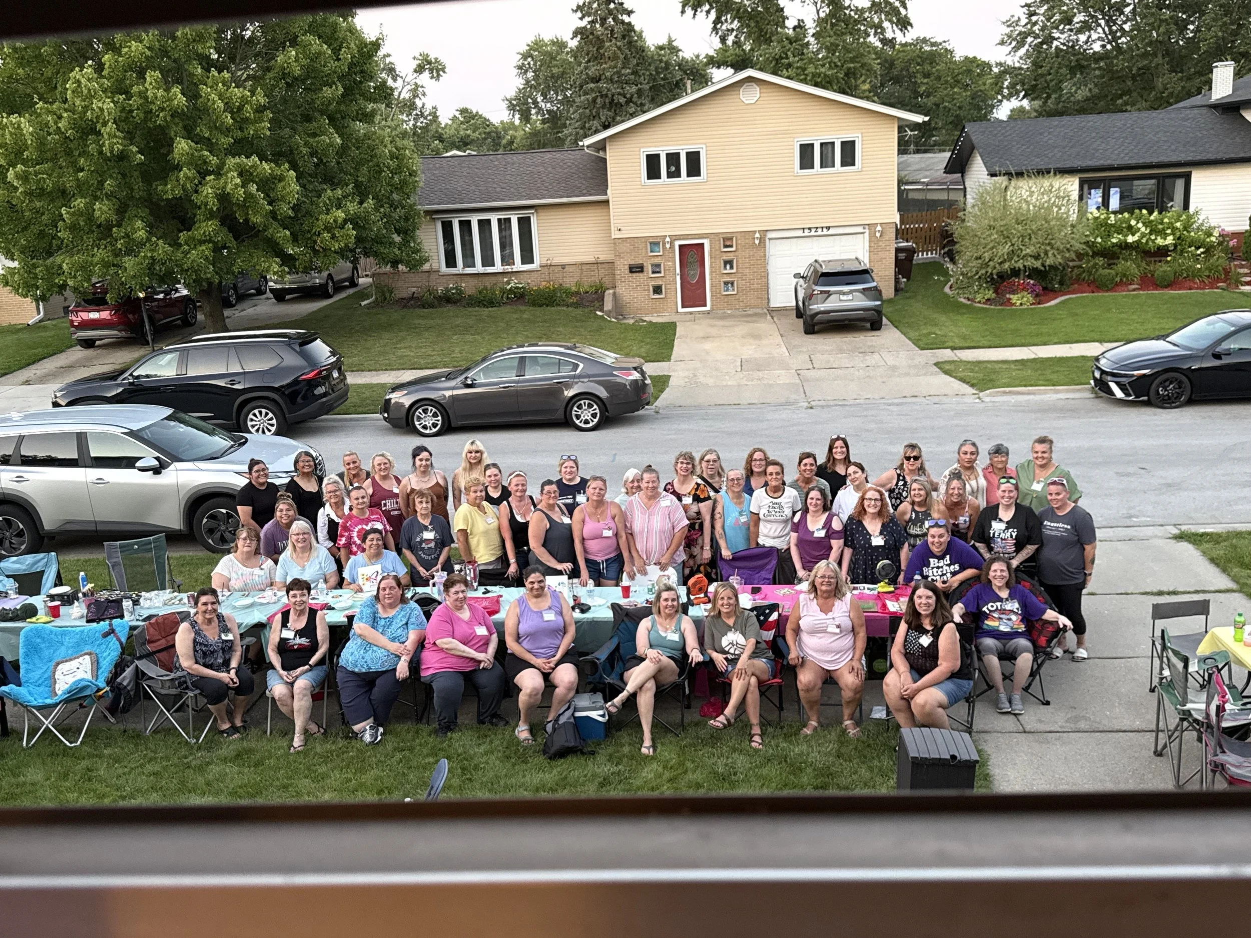 A large group of people is gathered outdoors in front of a suburban house with beige siding, a garage, and a landscaped yard, during daytime. Some people are sitting at tables with colorful tablecloths, and many are standing, all smiling and facing t