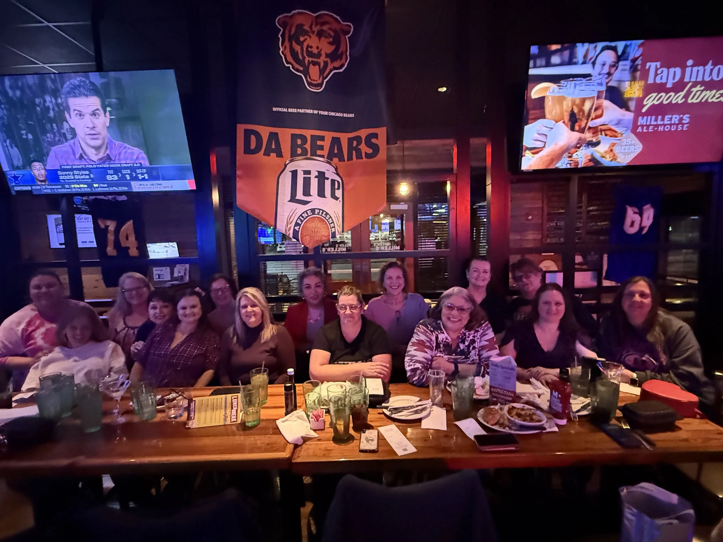 A group of people sitting at a restaurant table with plates of food and drinks, positioned in front of a large screen TV showing a news program and a colorful advertisement about Miller's ale-house, in a dimly lit restaurant.
