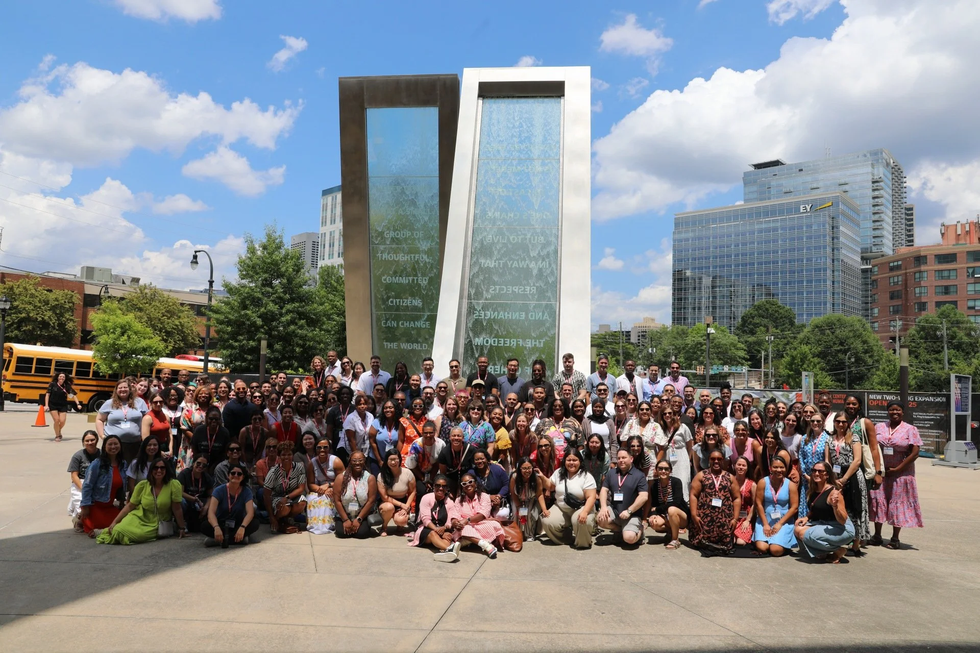 Large group of diverse people gathered outdoors in front of a large water feature sculpture with tall glass structures, city buildings, and a blue sky with clouds in the background.