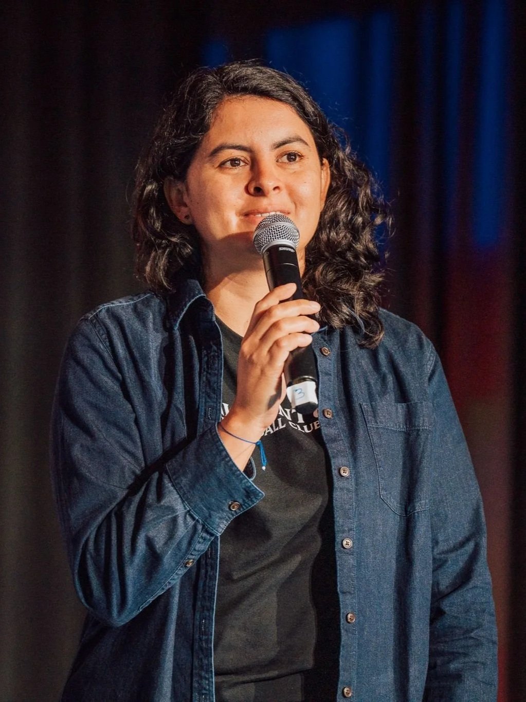 A woman with curly dark hair holding a microphone, standing in front of a dark background with red and blue curtains, wearing a denim shirt over a black T-shirt.