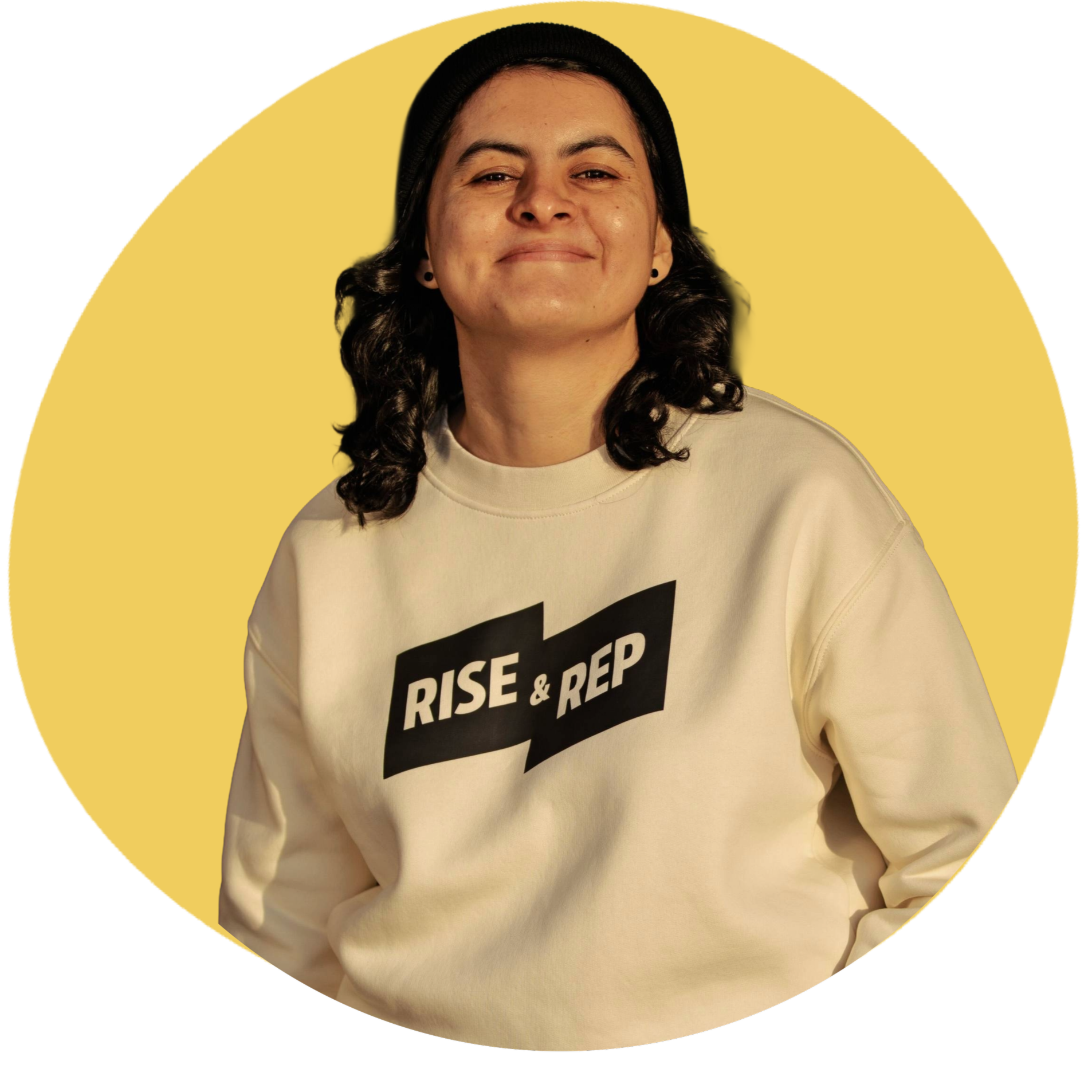 A woman with dark hair, wearing a black beanie and a beige sweatshirt with bold black text, smiling in front of a yellow background.