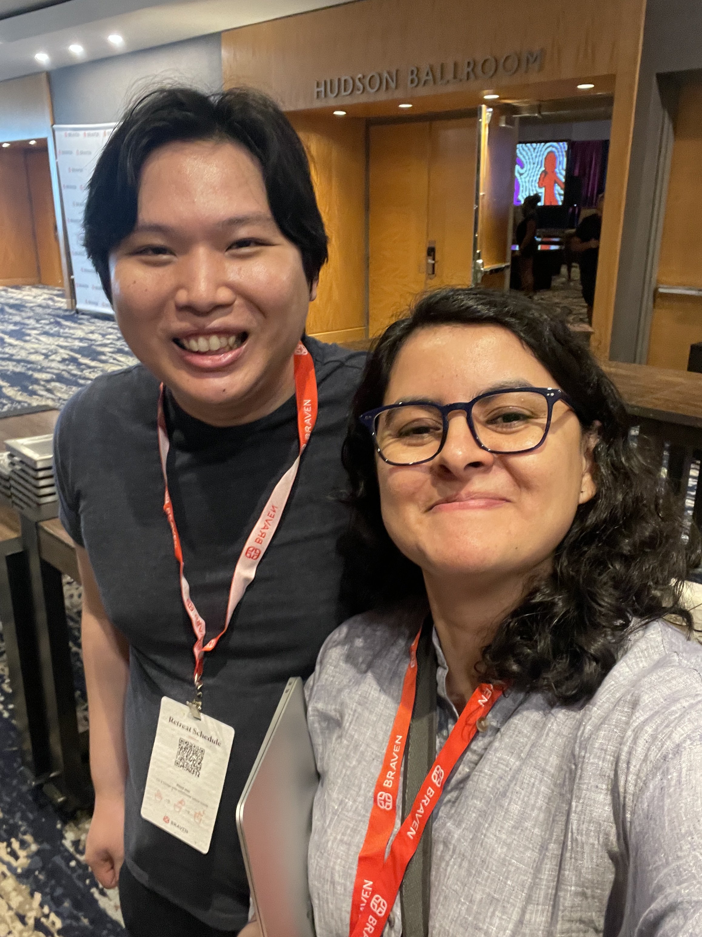 Two smiling women taking a selfie in a ballroom with a sign that reads 'Hudson Ballroom'. One woman has dark hair and glasses, the other has black hair and is wearing a gray blazer. Both are wearing orange lanyards.
