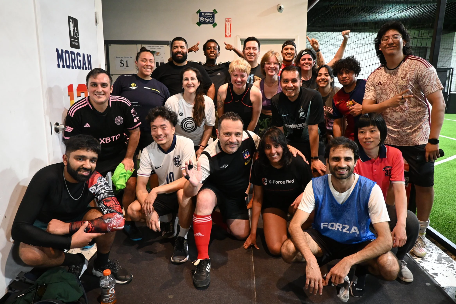 Group of diverse people posing together inside an indoor sports facility. They are smiling and making cheerful gestures, with some wearing sports jerseys and athletic clothing.