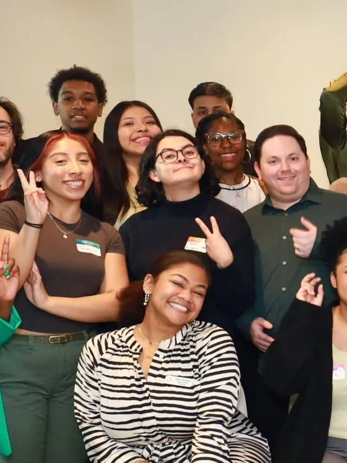 Group of smiling young adults, some making peace signs, at an indoor gathering or event.