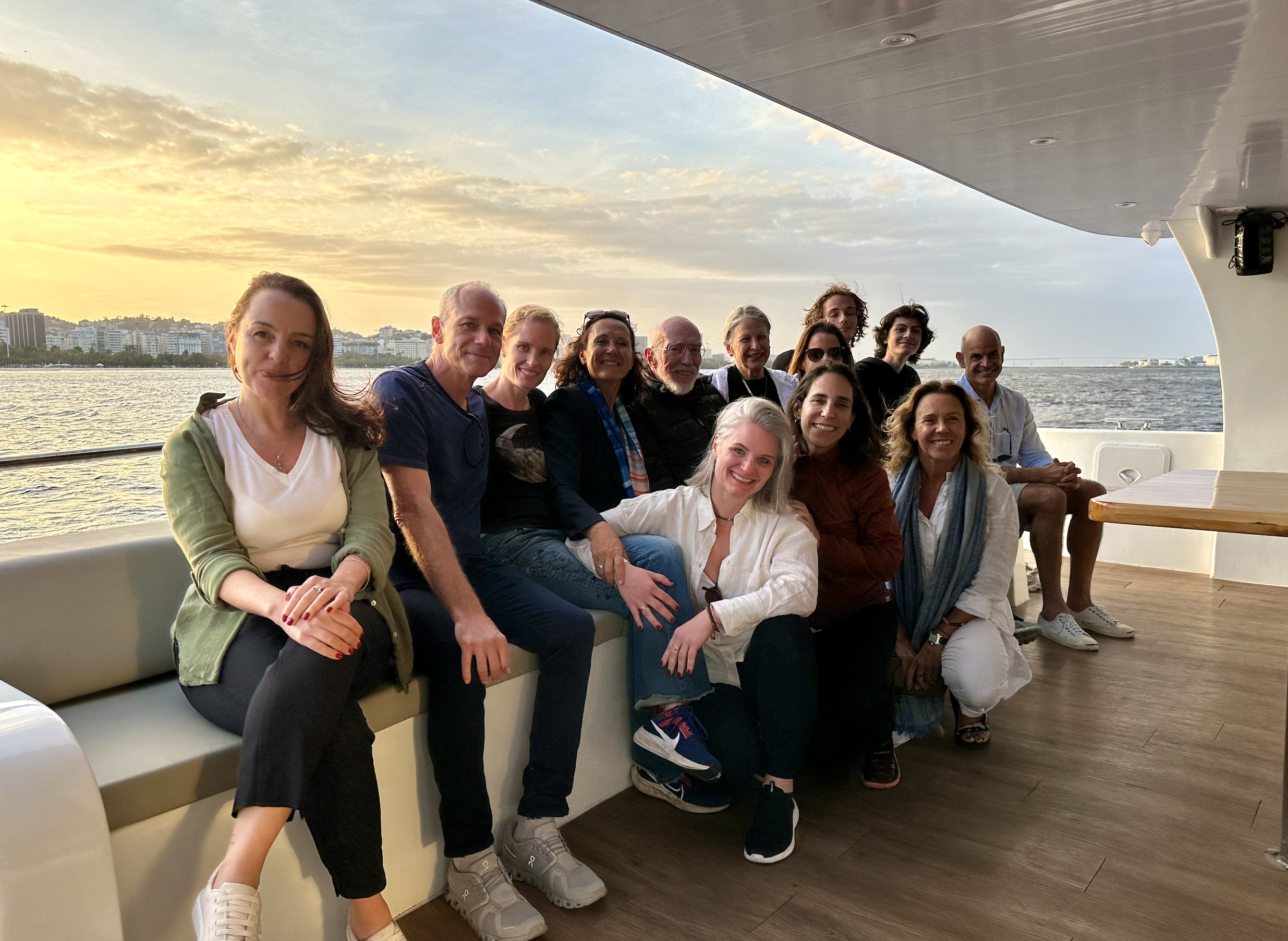 Grupo de pessoas sorrindo e posando para uma foto a bordo de um barco, com o rio e edifícios ao fundo, durante o pôr do sol.