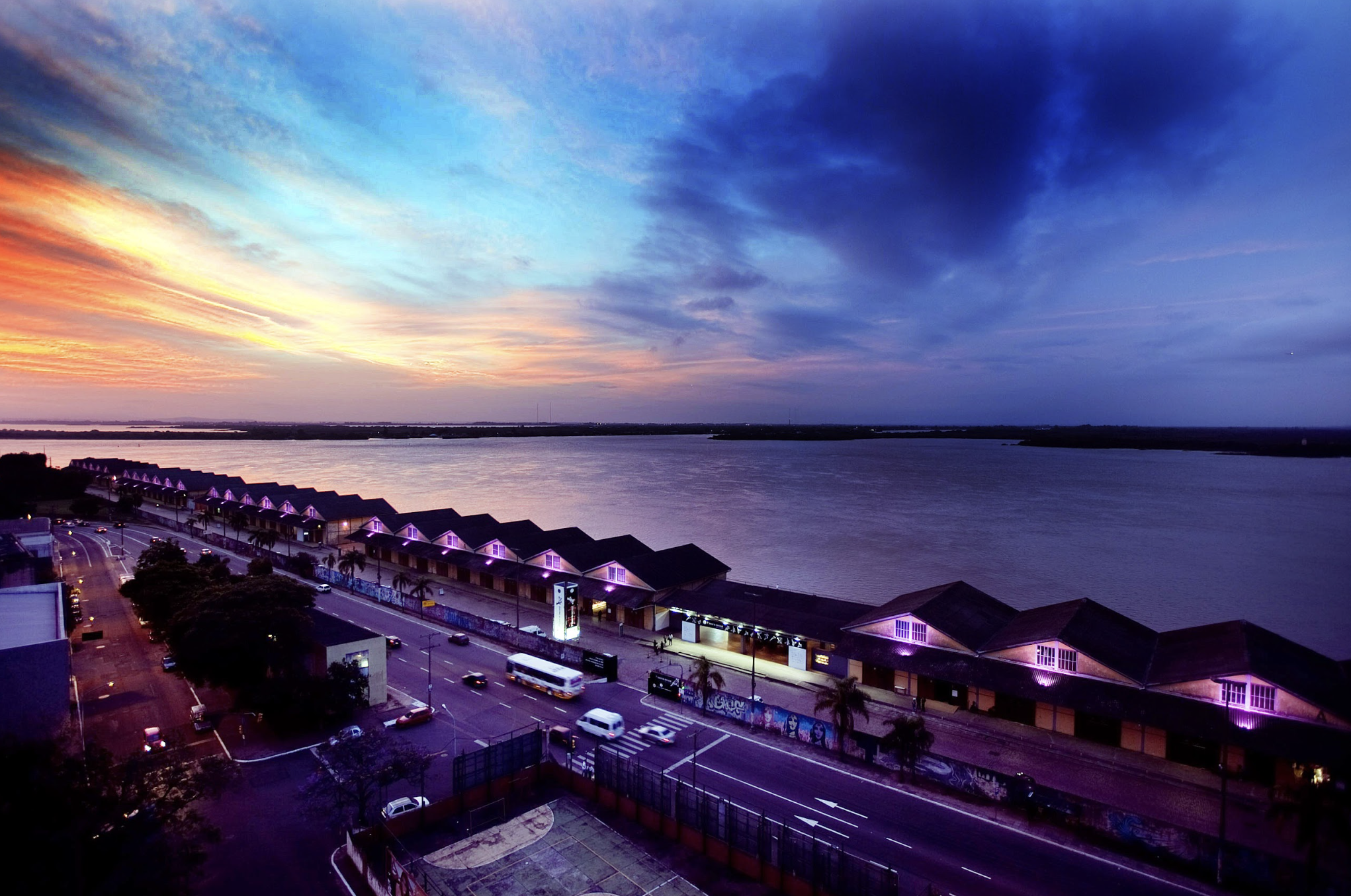 Cena de um pôr do sol com céu azul, laranja e roxo refletido na água de um rio ou lago, com prédios e casas ao longo da margem, além de uma rua movimentada com carros, e iluminação em toda a cena.