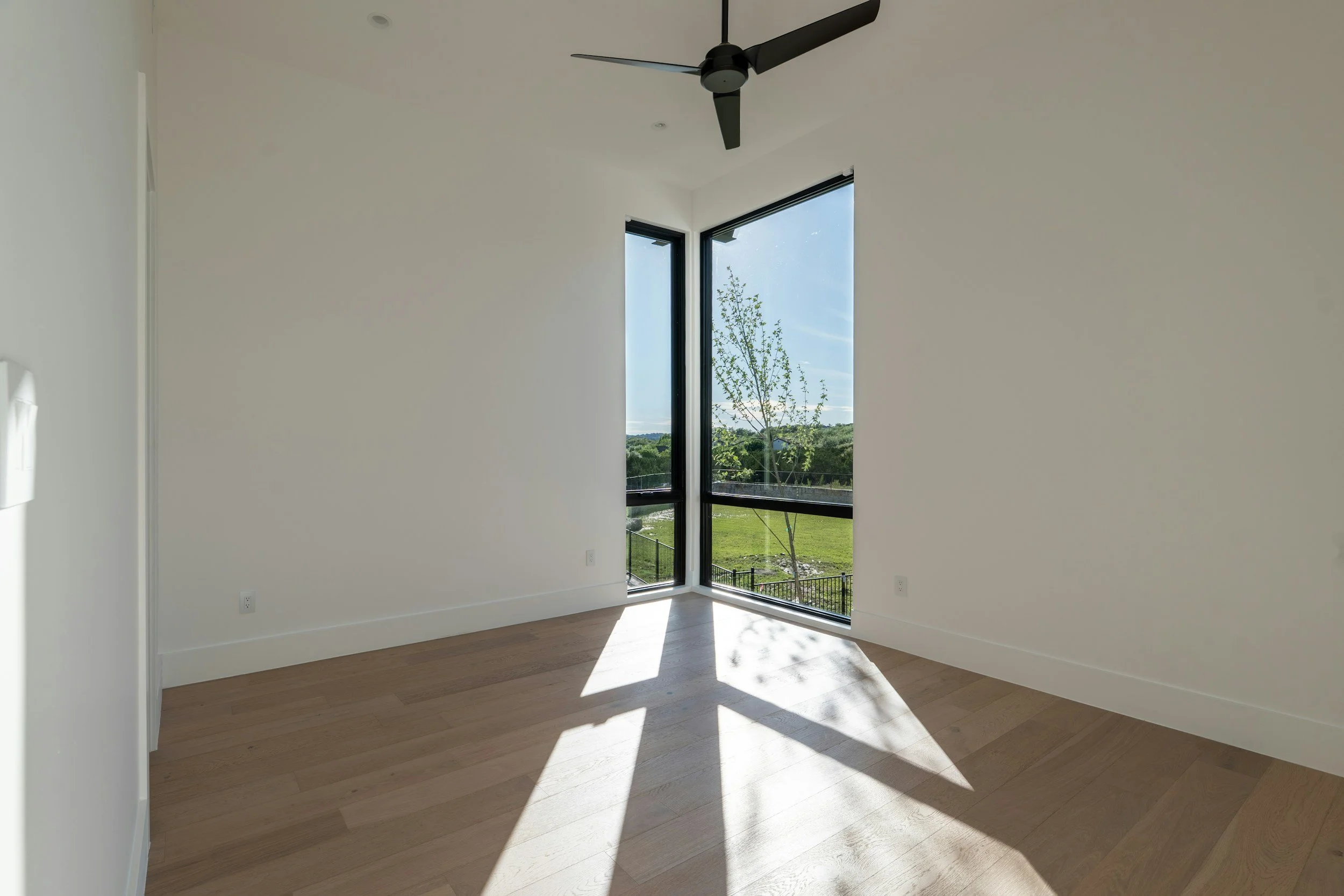 Empty room with large corner window showing outside trees and a grassy yard, sunlight casting shadows on light wood floor, white walls, and a modern black ceiling fan.