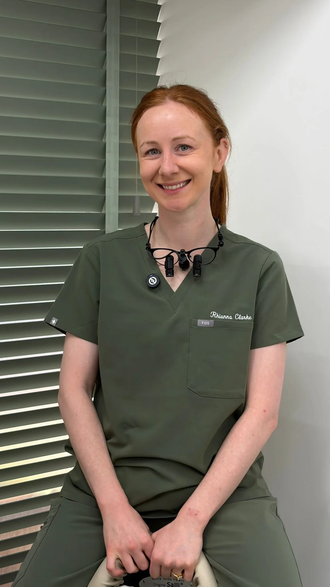 A smiling woman with red hair wearing green medical scrubs, a black lanyard, and magnifying glasses around her neck, sitting against a background with green blinds and white wall.