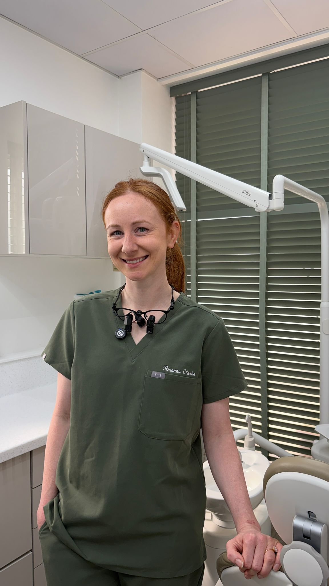 A smiling female healthcare worker wearing dark green scrubs and a name tag standing in a dental examination room, with dental equipment and a dental chair in the background.