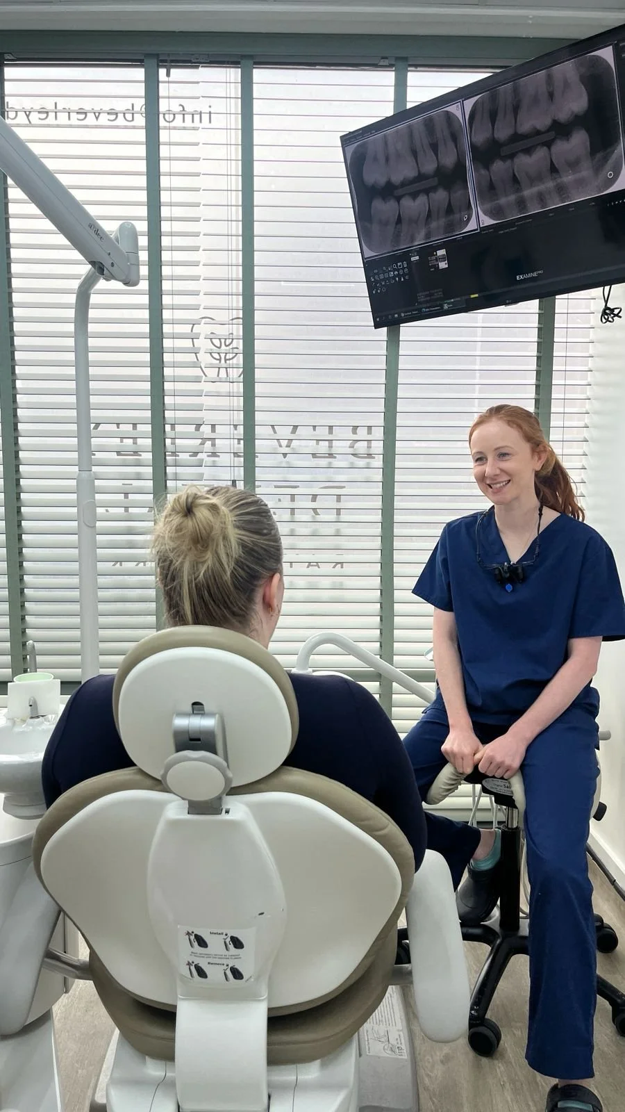 A dentist or dental hygienist in blue scrubs smiling and talking to a patient seated in a dental chair. The patient has blonde hair tied back and is viewed from behind. There are dental tools and a monitor displaying dental X-rays in the room with blinds and windows in the background.