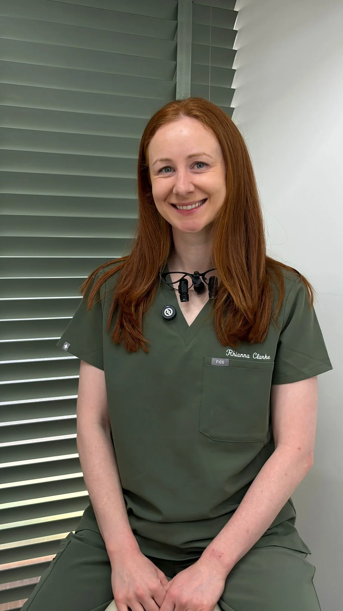 A woman with long red hair wearing green medical scrubs and a name tag that reads "Rhianna Clarke". She has a smile and is standing in front of green window shutters and a white wall, wearing a black lanyard with a medical device around her neck.