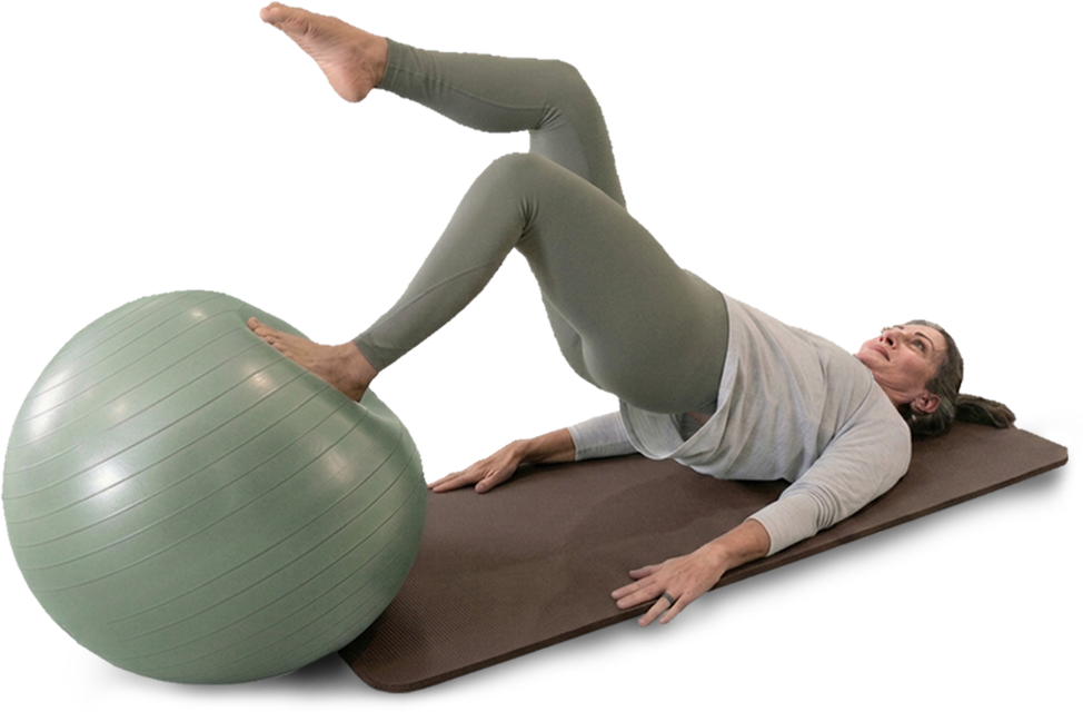 An older woman exercising on a brown yoga mat, lying with her shoulders on the mat, one leg raised and one leg positioned on a large green exercise ball, creating a single leg glute raise,  wearing green leggings and a light gray long-sleeve shirt.